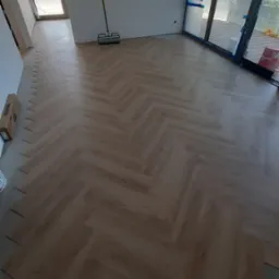 Ułożony parkiet w jodełkę, jasne drewno, widoczne narzędzie do montażu, okno zabezpieczone folią malarską. Częściowo ułożone panele w rogu pomieszczenia.