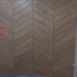 BUD VAS - Ściana wykończona drewnopodobnymi panelami ułożonymi we wzór jodełki; widoczne fugi i fragmenty taśmy malarskiej. Ciekawy design.