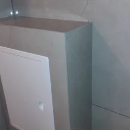 Szara zabudowa z płyt imitujących marmur, ukrywająca instalacje w łazience, z białą klapą rewizyjną z okrągłym uchwytem. Minimalistyczny design.