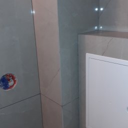BUD VAS - Wykończona ściana w łazience z szarych płytek imitujących marmur, widoczny otwór na baterię i biała klapa rewizyjna. Subtelne oświetlenie półki.
