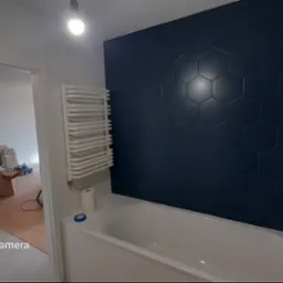 Nowoczesna łazienka w trakcie wykończenia: ciemne, heksagonalne płytki na ścianie nad wanną, biały grzejnik drabinkowy i widok na remontowane pomieszczenie.