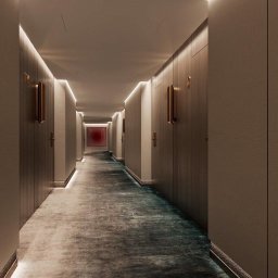 BUD VAS - Długi korytarz hotelowy z drewnianymi drzwiami, oświetleniem LED i wzorzystą wykładziną. W tle widoczny obraz w czerwonej ramie, tworzący eleganckie wnętrze.