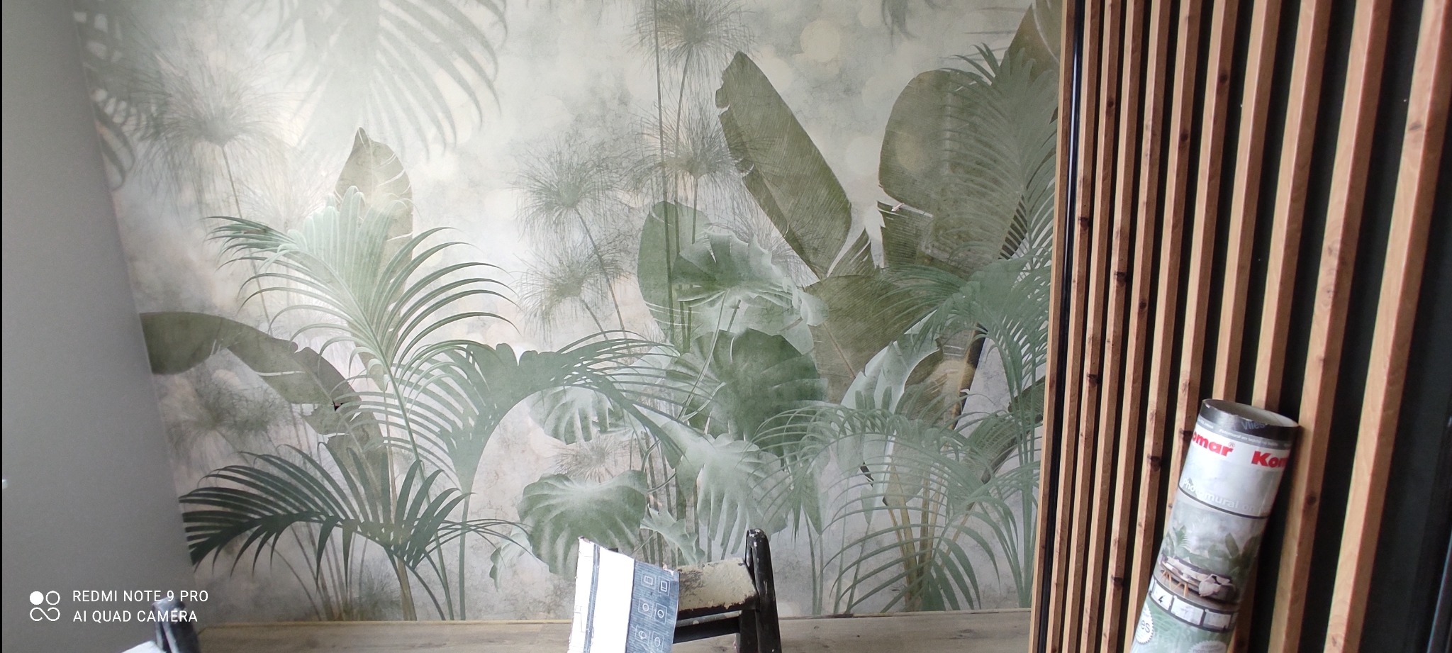 Ściana z fototapetą w motywy tropikalne, częściowo zasłonięta drewnianymi lamelami; widoczny fragment krzesła i rolka fototapety Komar.