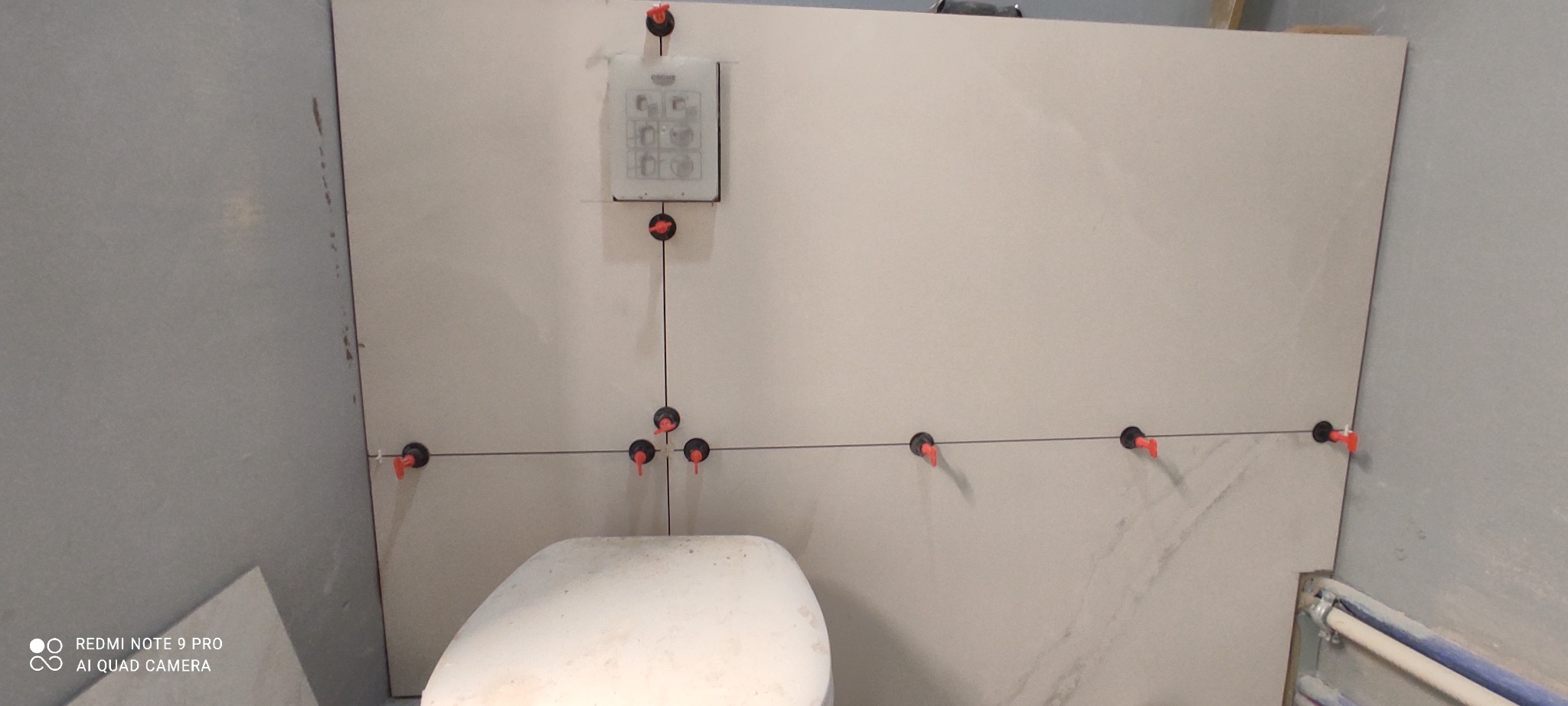 Świeżo ułożone, duże płytki ceramiczne na ścianie w łazience z widocznymi systemami poziomowania płytek i częściowo widoczną toaletą w dolnej części kadru.