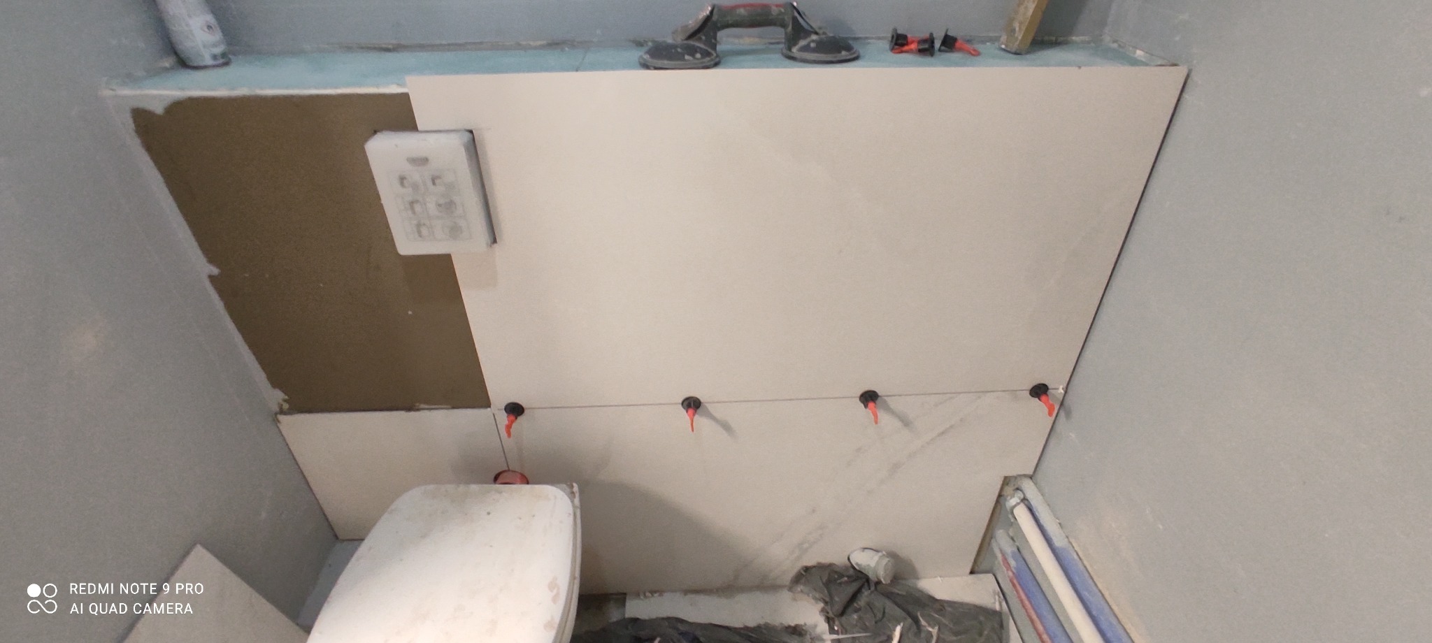 Montaż jasnych płytek w łazience z użyciem poziomowania, widoczne narzędzia glazurnicze i fragment toalety.