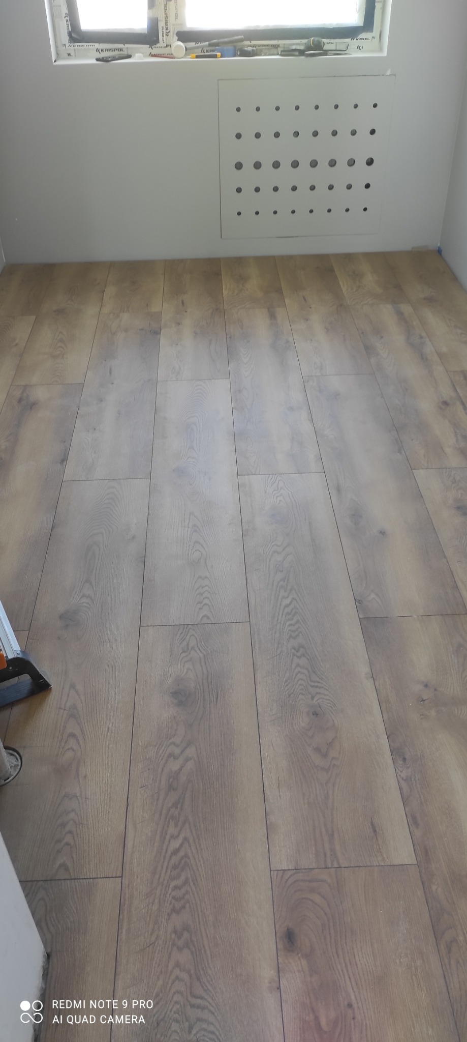 Ułożone panele podłogowe w jasnym odcieniu drewna, widoczny fragment narzędzia do montażu paneli przy ścianie, biała ściana z kratką wentylacyjną nad podłogą.