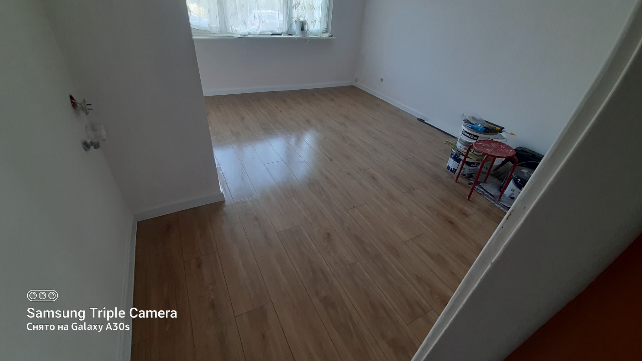 Jasne panele podłogowe w pokoju z białymi ścianami, widoczne puszki z farbą i czerwony taboret w rogu.