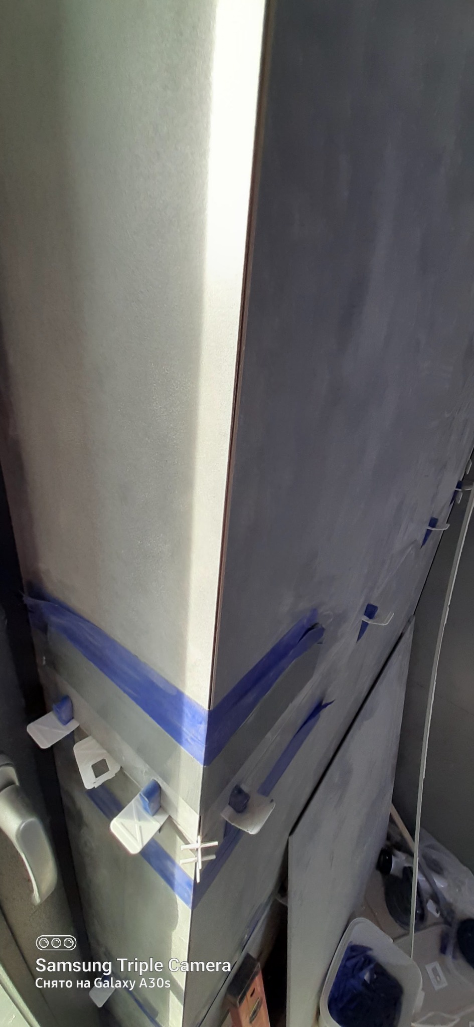 Układanie szarych płytek na narożniku ściany, zabezpieczonych niebieską taśmą i białymi krzyżykami dystansowymi, widoczne narzędzia i wiadro z gąbkami.