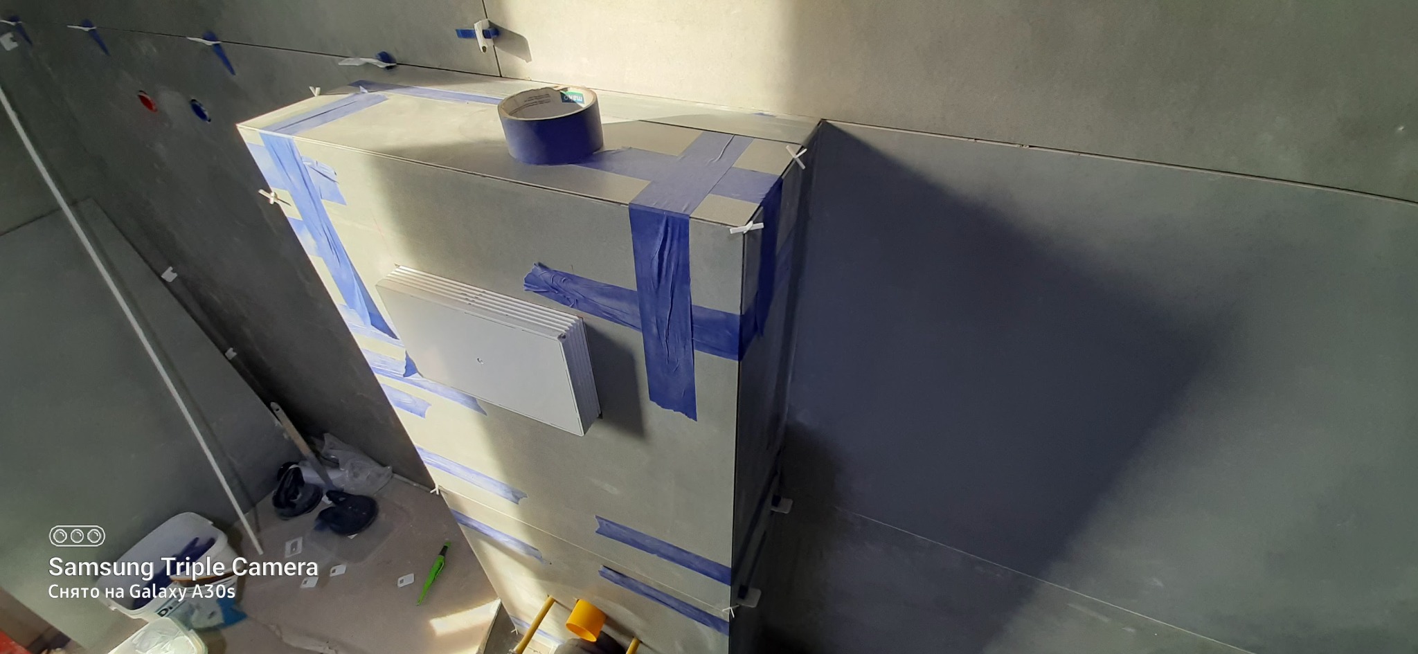 Szara ściana w trakcie układania płytek, zabezpieczona niebieską taśmą malarską i klinami dystansowymi, widoczny fragment wentylacji, ujęcie z dołu.