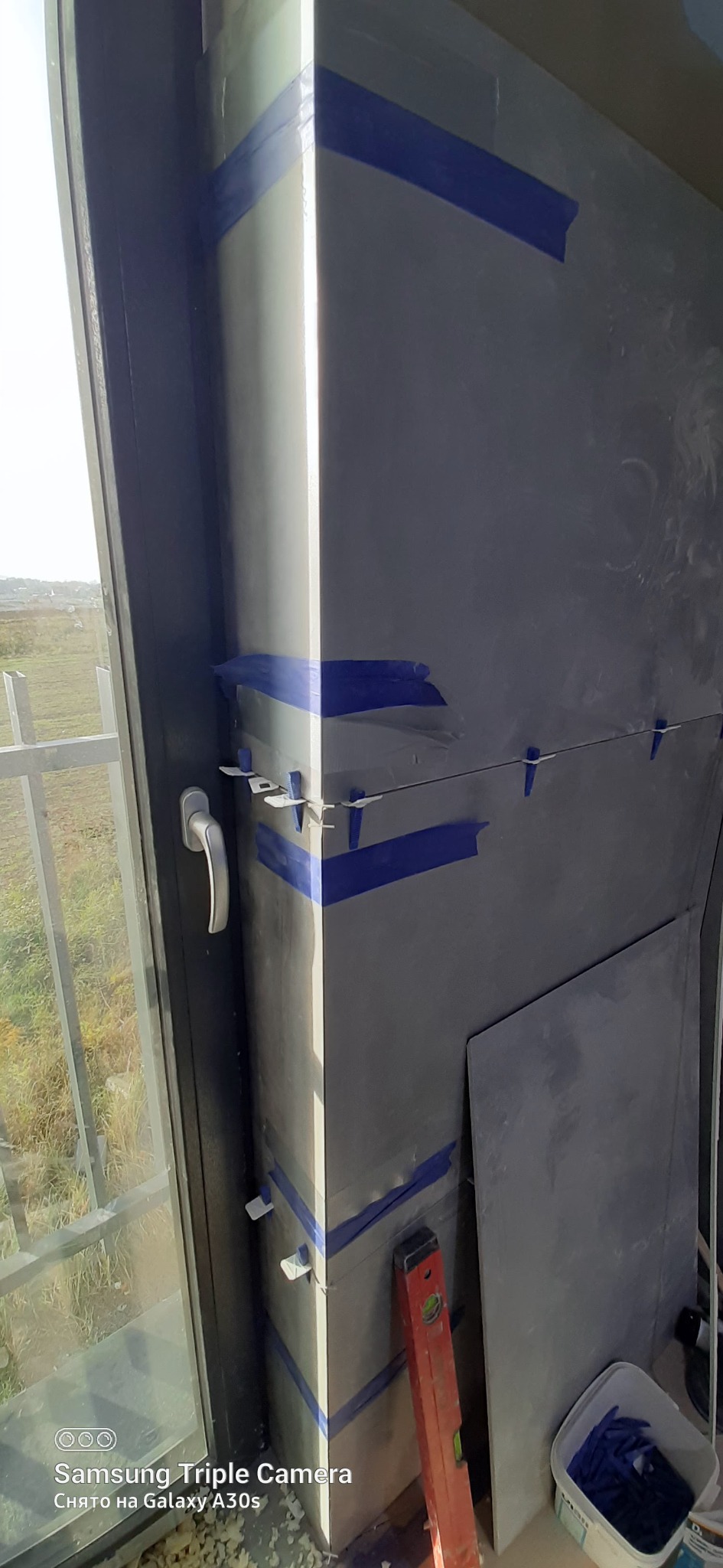 Szara ściana z płytami, zabezpieczona niebieską taśmą i plastikowymi krzyżykami dystansowymi, w tle okno z widokiem na zewnątrz, obok poziomica i wiaderko z krzyżykami.