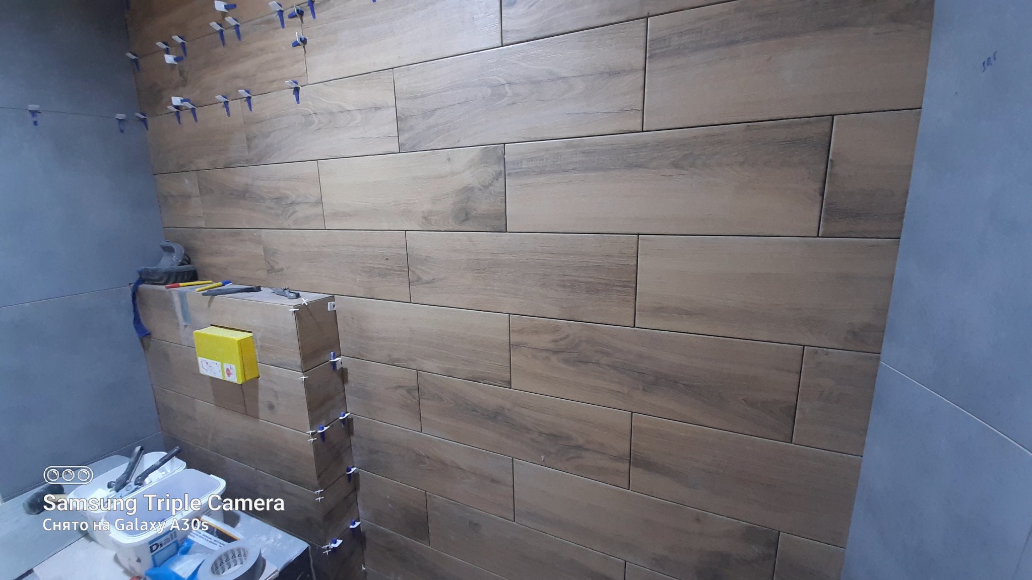 Ściana w trakcie wykańczania z ułożonymi płytkami imitującymi drewno, widoczne narzędzia glazurnicze i plastikowe krzyżyki dystansowe.