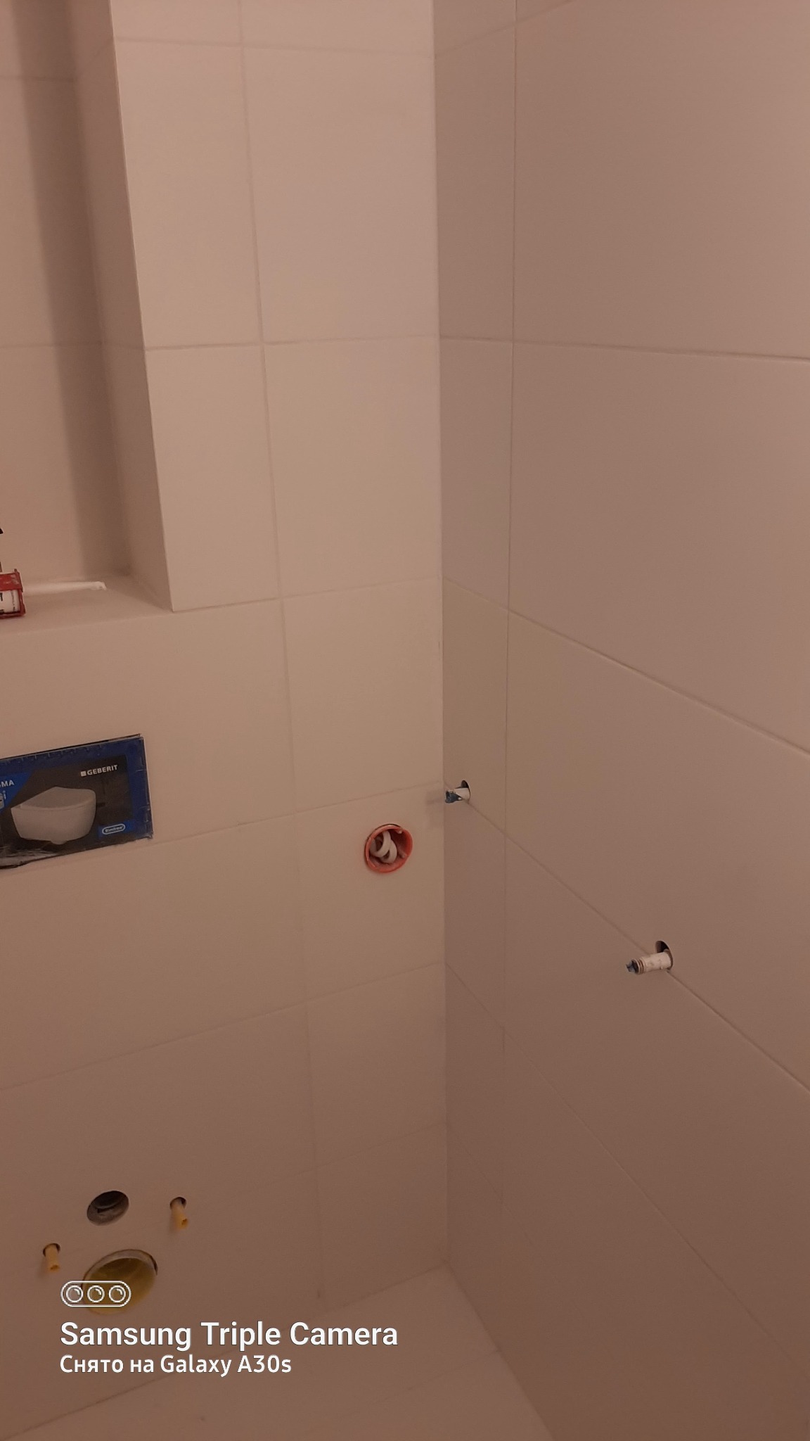 Wnętrze łazienki w trakcie wykańczania: białe płytki na ścianach, przygotowane punkty instalacyjne, widoczna puszka podtynkowego systemu spłukiwania Geberit.