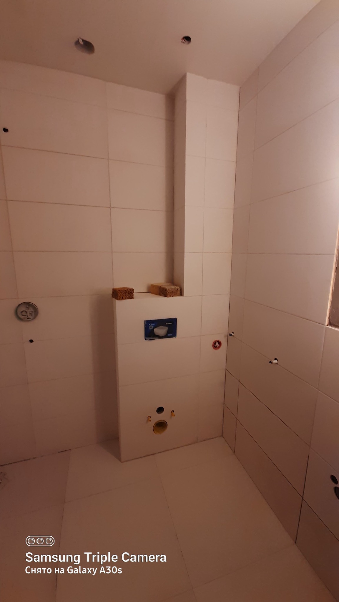 Wnętrze łazienki w trakcie wykańczania, widoczne białe płytki, zabudowany stelaż WC z zamontowanym przyciskiem spłukującym, dwa gąbki na stelażu i przygotowane otwory na armaturę.