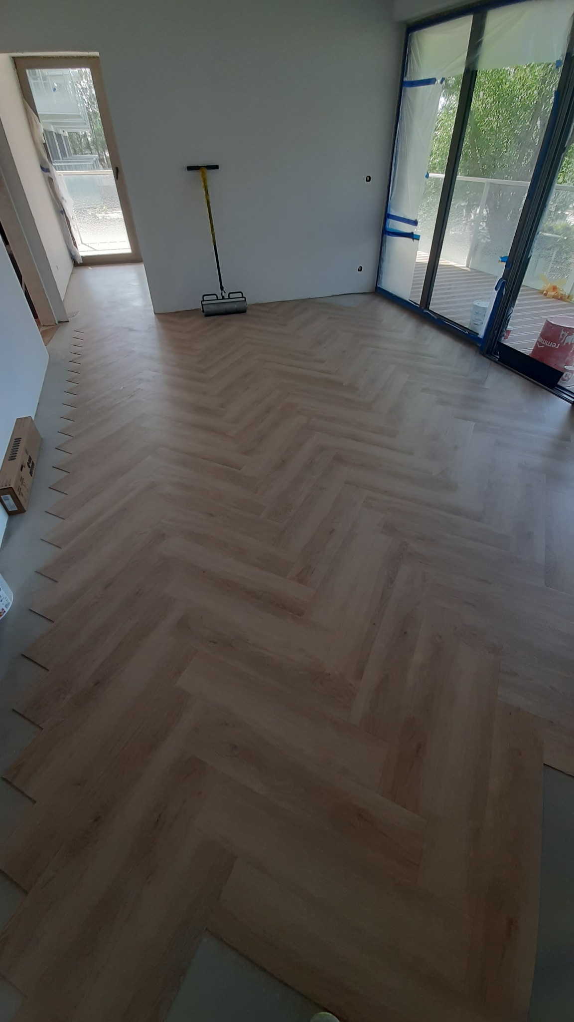 Ułożony parkiet w jodełkę, jasne drewno, widoczne narzędzie do montażu, okno zabezpieczone folią malarską. Częściowo ułożone panele w rogu pomieszczenia.