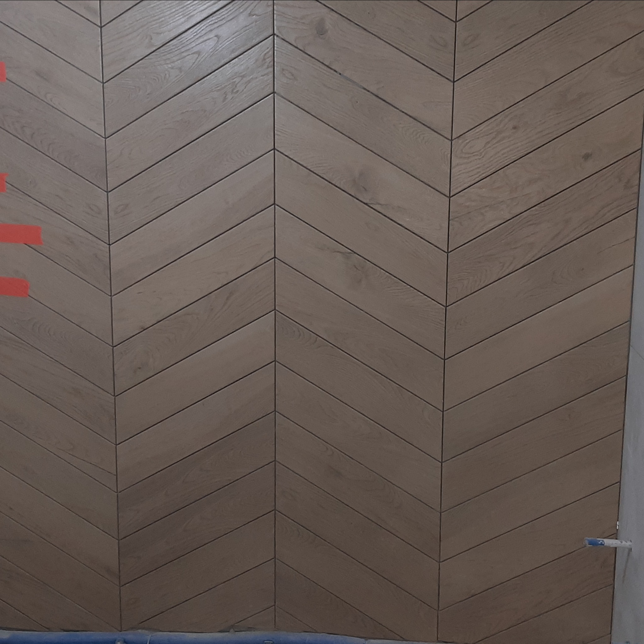 Ściana wykończona drewnopodobnymi panelami ułożonymi we wzór jodełki; widoczne fugi i fragmenty taśmy malarskiej. Ciekawy design.