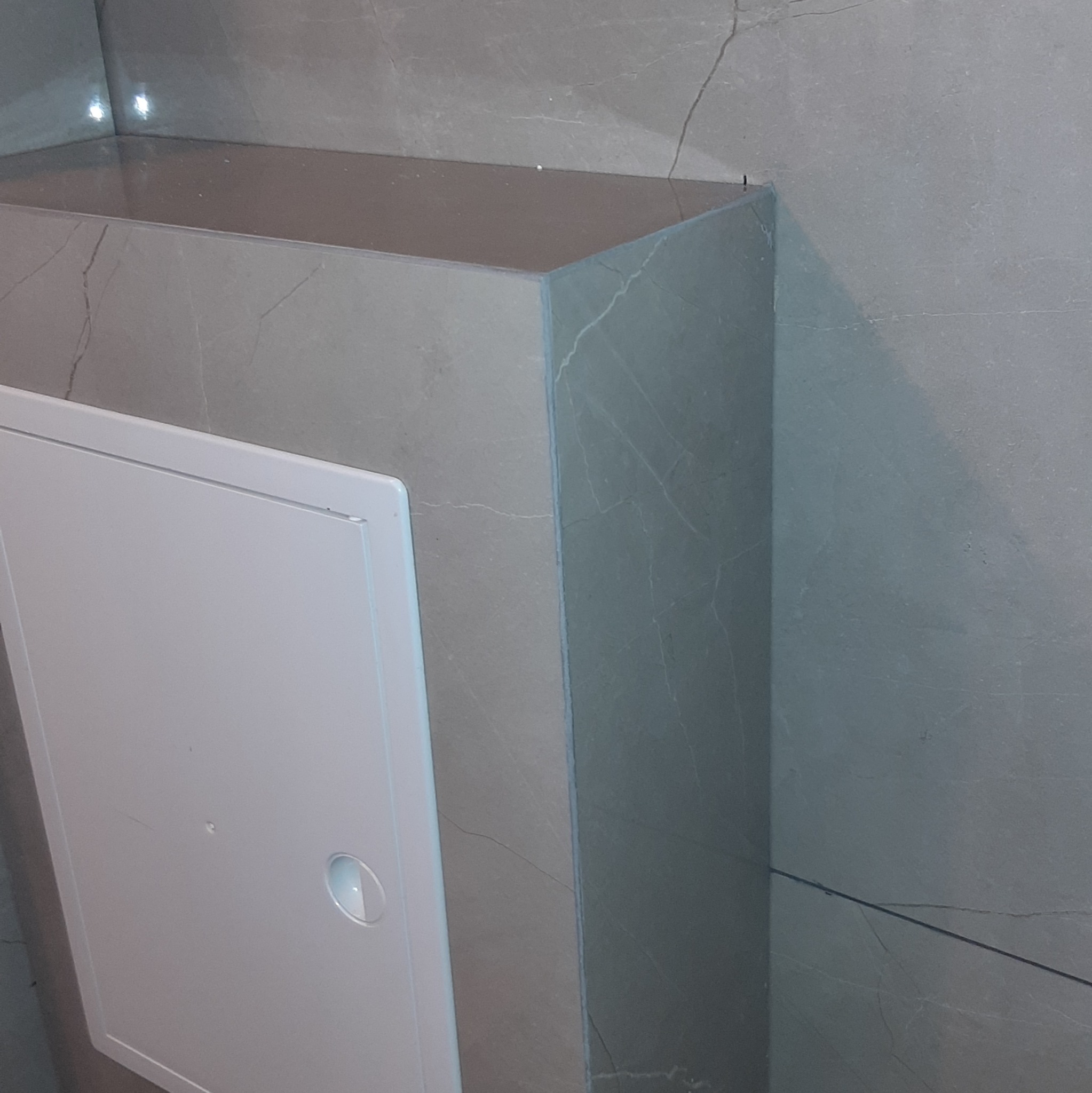 Szara zabudowa z płyt imitujących marmur, ukrywająca instalacje w łazience, z białą klapą rewizyjną z okrągłym uchwytem. Minimalistyczny design.