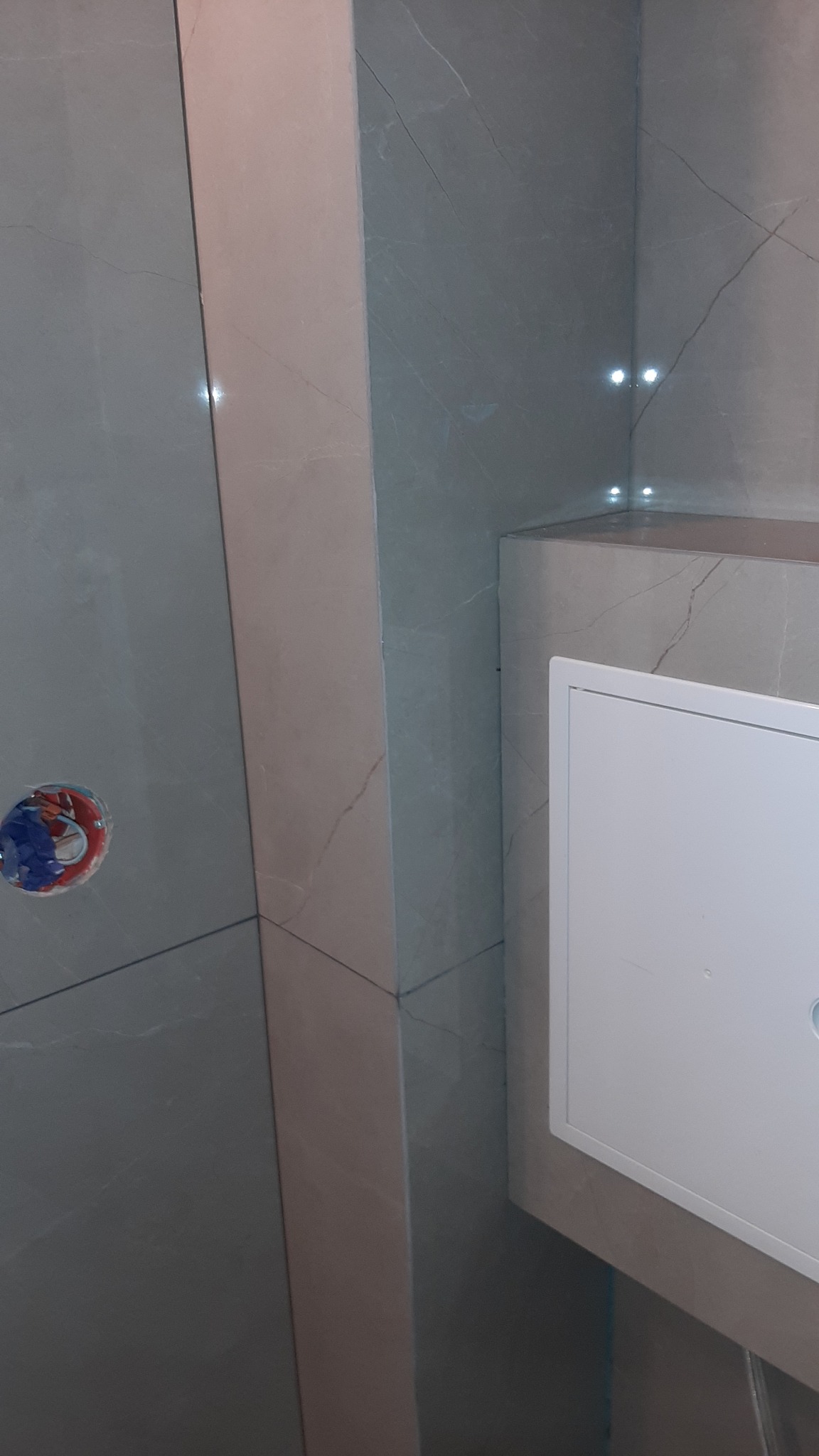 Wykończona ściana w łazience z szarych płytek imitujących marmur, widoczny otwór na baterię i biała klapa rewizyjna. Subtelne oświetlenie półki.