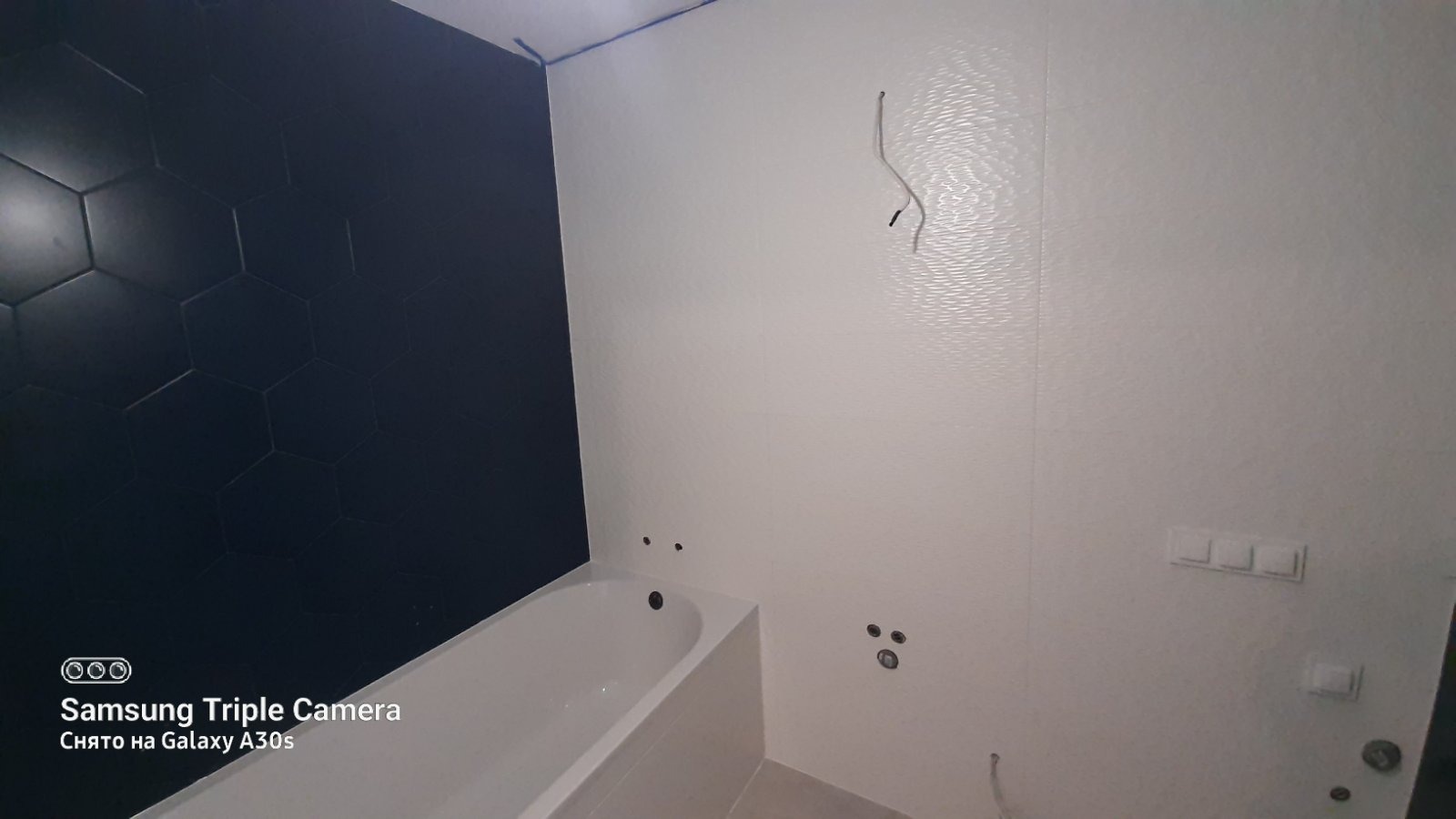 Remont łazienki: widok na wannę, białe i czarne heksagonalne płytki na ścianach, przygotowane punkty instalacyjne. Widoczne niedociągnięcia wykończeniowe.