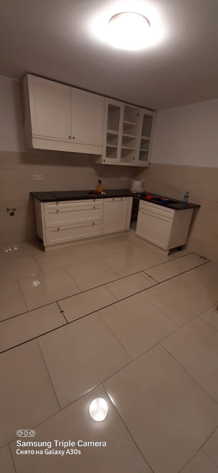 Jasna kuchnia po remoncie: kremowe szafki z czarnym blatem, beżowe płytki podłogowe z odpływem liniowym. Widok na całe pomieszczenie, ukończone prace.