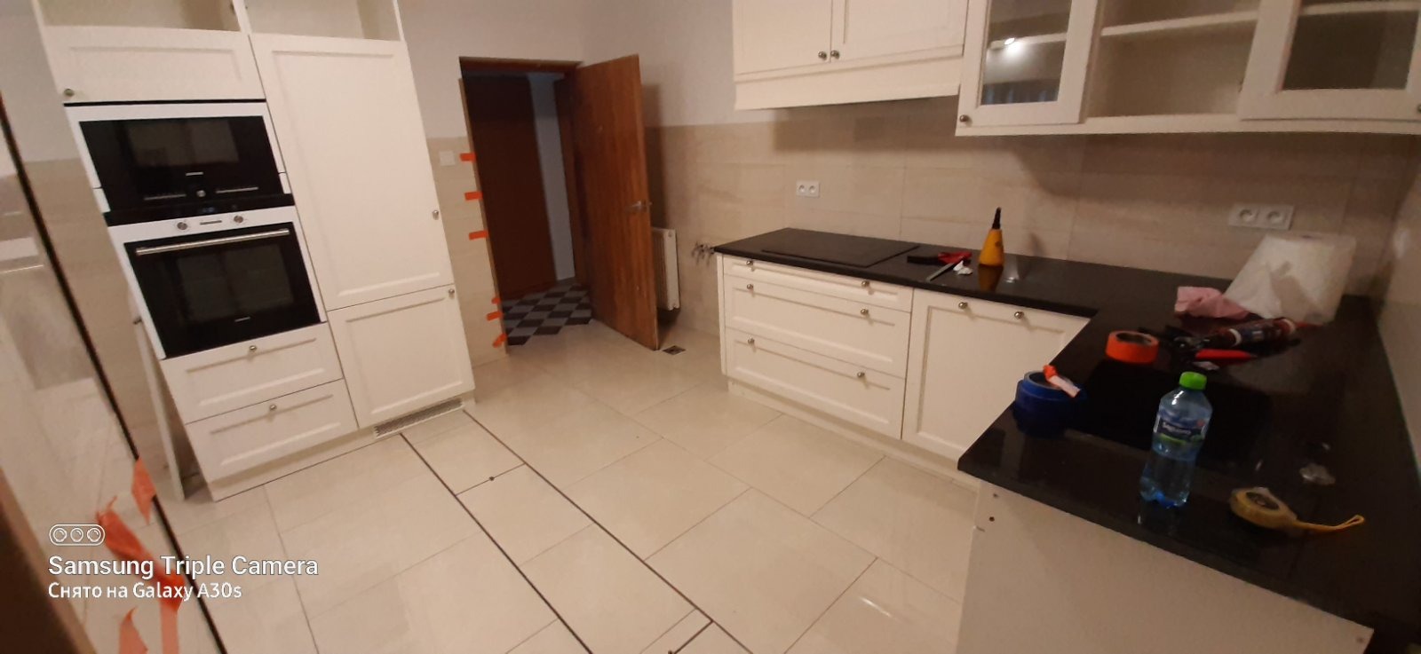 Jasna kuchnia po remoncie: białe szafki, czarny blat, płytki podłogowe. Widoczne narzędzia i akcesoria na blacie. Otwarta drewniana  do innego pomieszczenia.