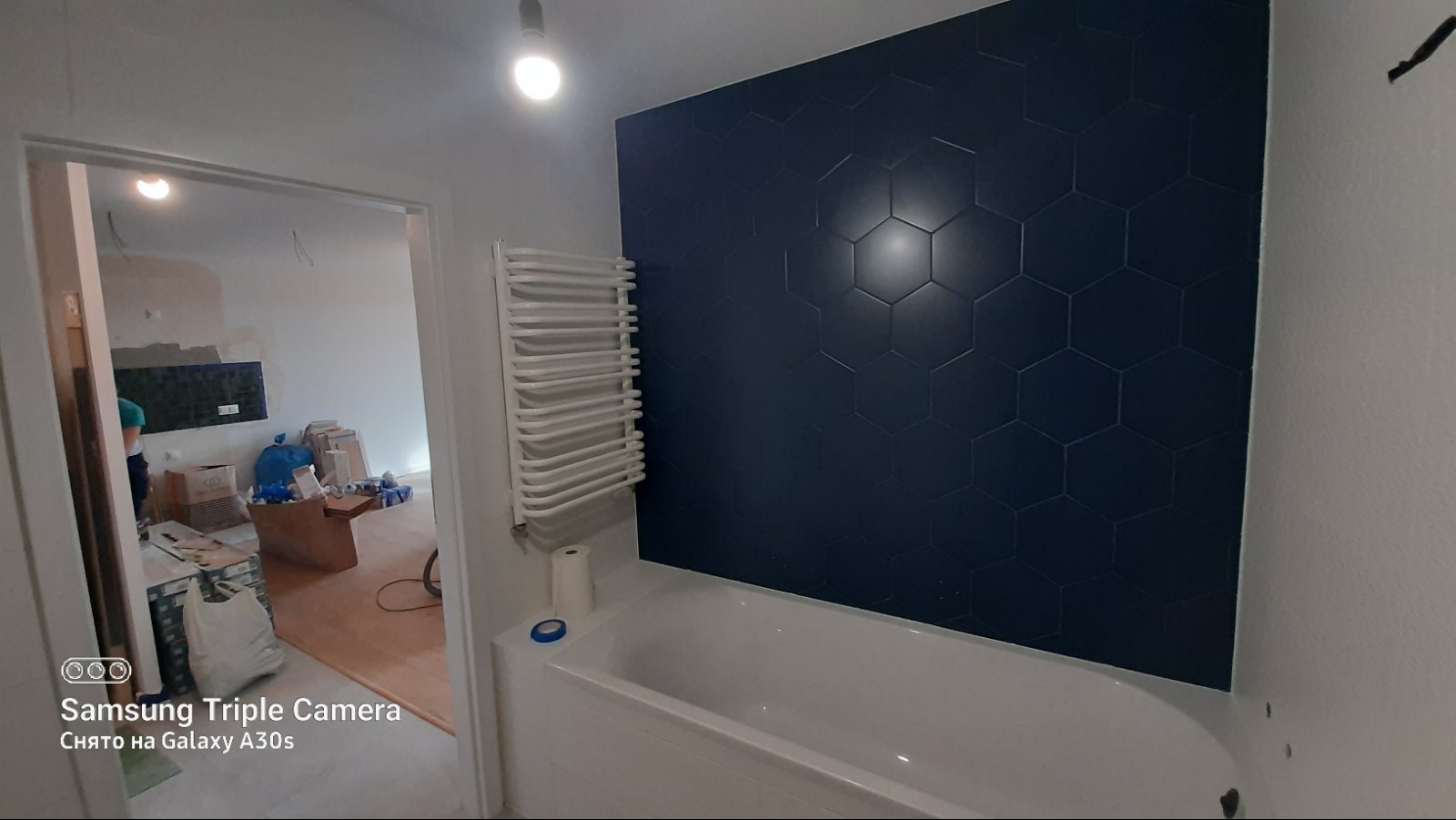 Nowoczesna łazienka w trakcie wykończenia: ciemne, heksagonalne płytki na ścianie nad wanną, biały grzejnik drabinkowy i widok na remontowane pomieszczenie.