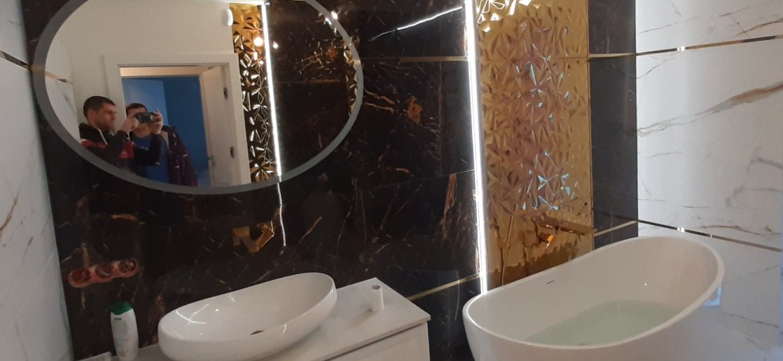 Elegancka łazienka z owalnym lustrem, marmurowymi płytkami w odcieniach bieli i czerni, złotymi akcentami oraz wolnostojącą wanną wypełnioną wodą.