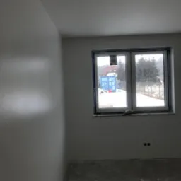 Świeżo wyszpachlowane, białe ściany w pustym pokoju z widokiem na zimowy krajobraz przez okno. Widoczne ślady po szpachlowaniu i dwa gniazdka elektryczne.