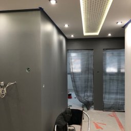 WOJ-PUTZ HUBERT WOJACZEK - Szare ściany w trakcie malowania, zabezpieczone taśmą malarską przy suficie, perforowany panel oświetleniowy na suficie, zakryte folią drzwi w tle, krzesło z narzędziami na pierwszym planie.