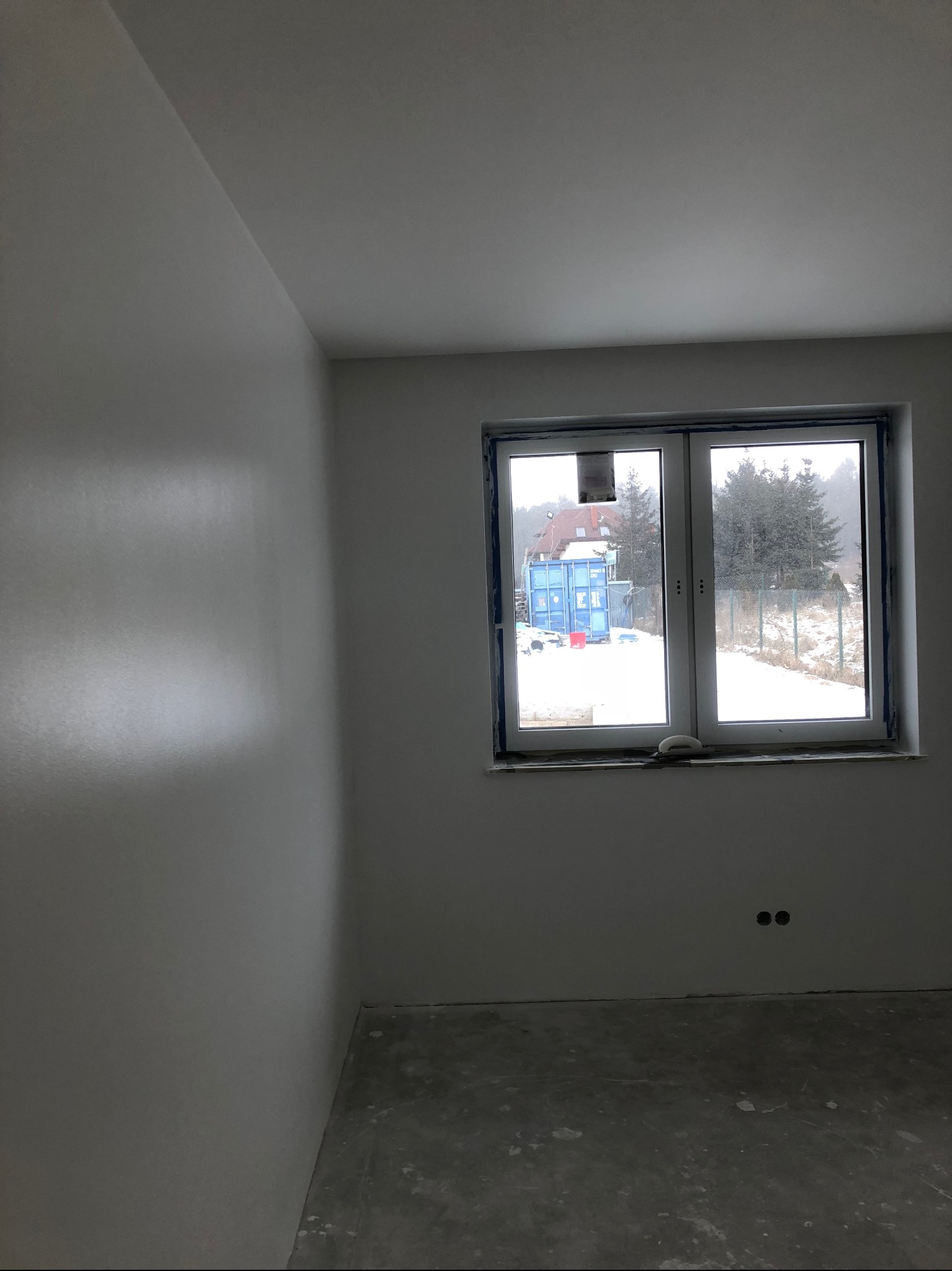 Świeżo wyszpachlowane, białe ściany w pustym pokoju z widokiem na zimowy krajobraz przez okno. Widoczne ślady po szpachlowaniu i dwa gniazdka elektryczne.