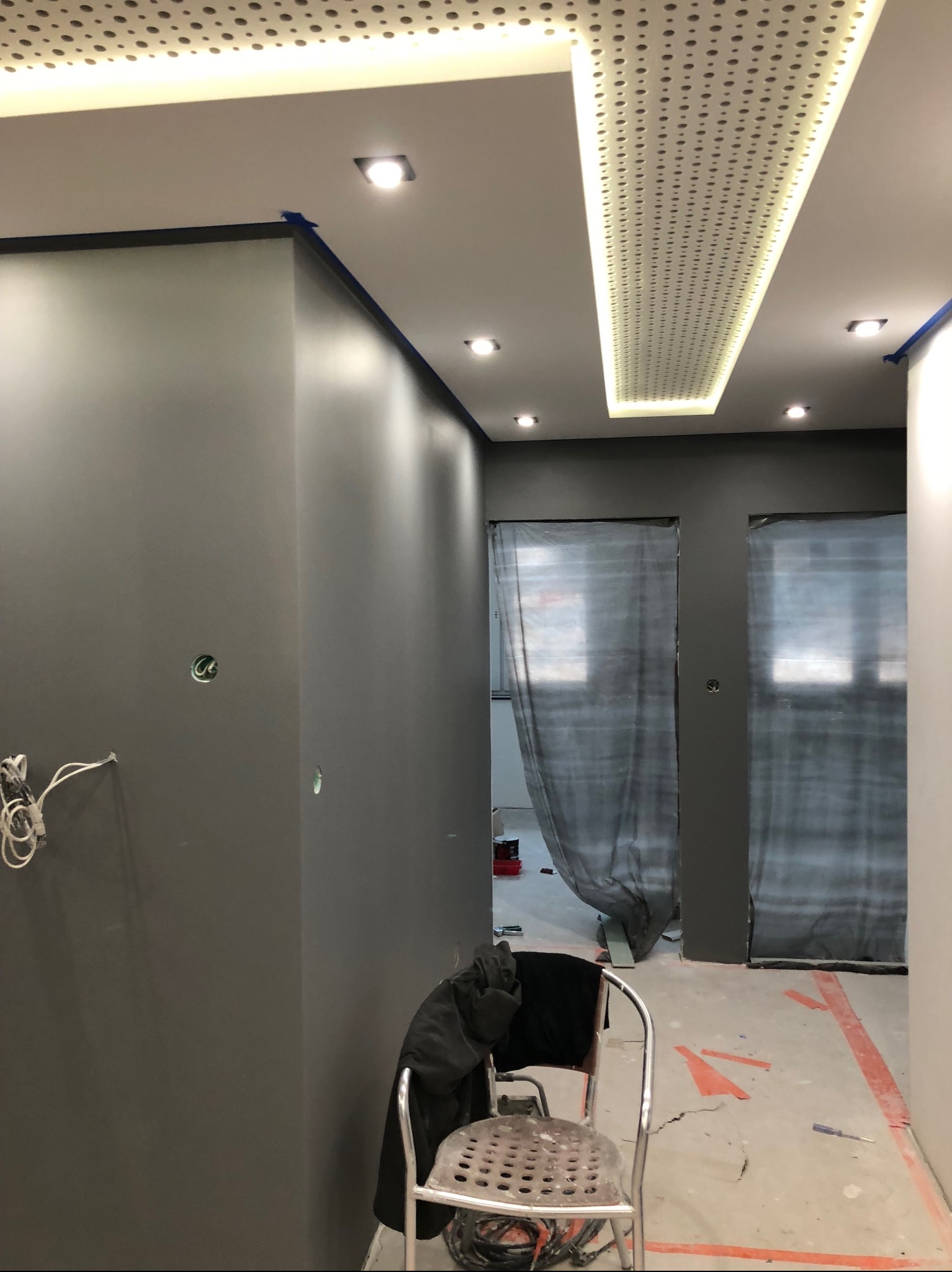 Szare ściany w trakcie malowania, zabezpieczone taśmą malarską przy suficie, perforowany panel oświetleniowy na suficie, zakryte folią drzwi w tle, krzesło z narzędziami na pierwszym planie.
