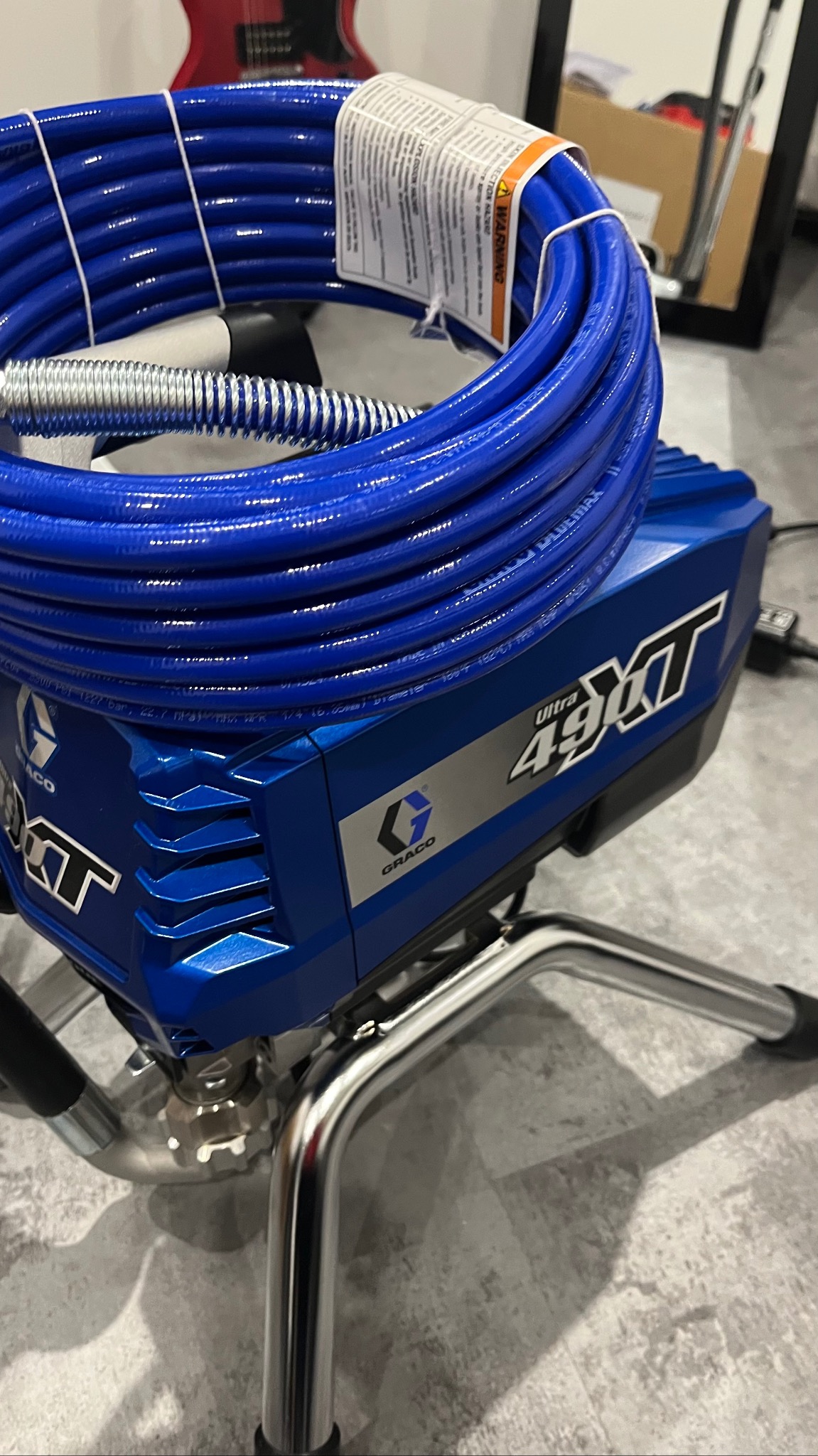 Niebieski agregat malarski Graco Ultra 490XT z nawiniętym wężem i instrukcją, stojący na chromowanym stelażu.