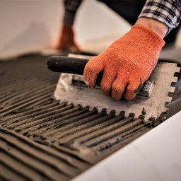 Perfect Home Poland - Nakładanie zaprawy klejowej zębatą pacą na podłoże pod płytki, pomarańczowa rękawica robocza, wzór w rowki, widoczne buty robocze w tle.