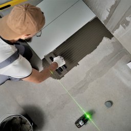 Perfect Home Poland - Układanie jasnych płytek podłogowych z użyciem poziomicy laserowej, widoczna paca z klejem oraz wiadro z zaprawą.