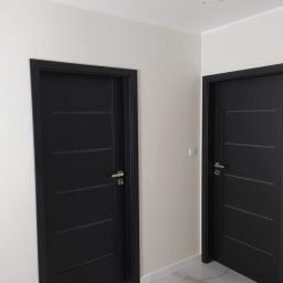 Perfect Home Poland - Dwa czarne, nowoczesne drzwi wewnętrzne z poziomymi frezami, zamontowane w jasnym pomieszczeniu z marmurową podłogą i białymi ścianami. Widoczne wystające kable ze ściany w miejscu lampy.