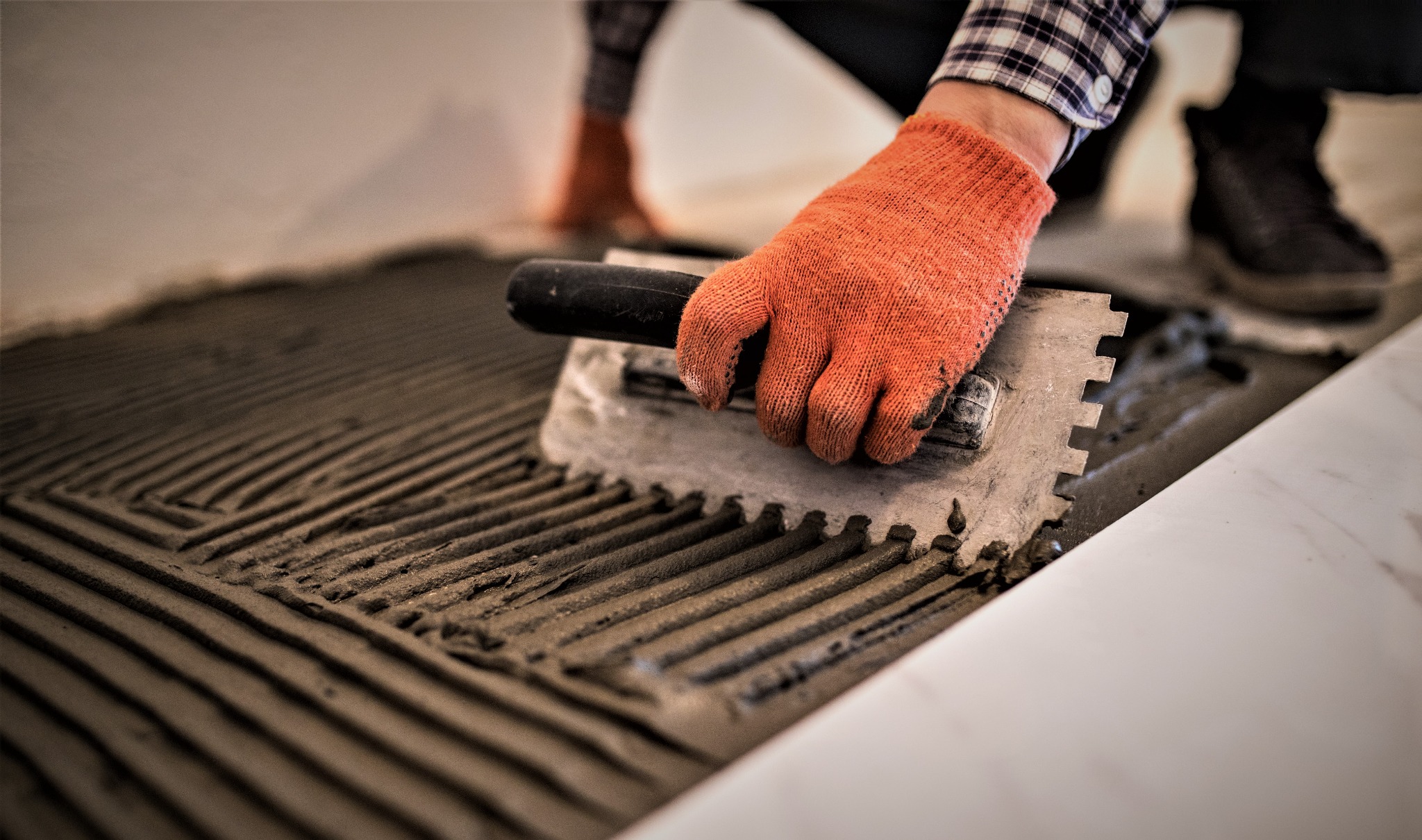 Nakładanie zaprawy klejowej zębatą pacą na podłoże pod płytki, pomarańczowa rękawica robocza, wzór w rowki, widoczne buty robocze w tle.