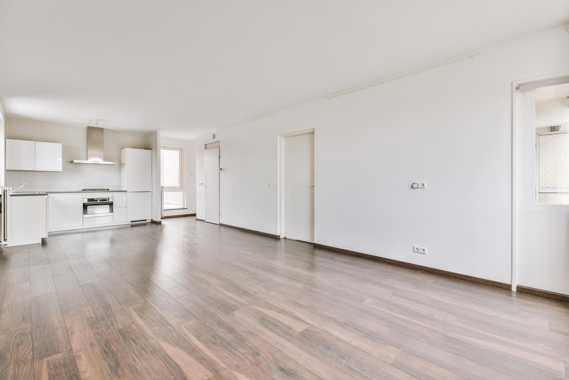 Jasne, przestronne wnętrze mieszkania z drewnopodobną podłogą, białą kuchnią i ścianami, widoczne gniazdka elektryczne na ścianie.