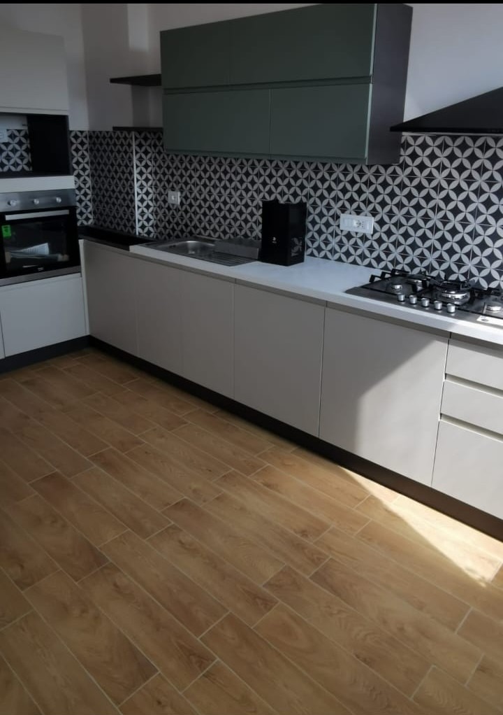 Nowoczesna kuchnia w odcieniach szarości i drewna z geometrycznym wzorem na ścianie, widok na zabudowę kuchenną z blatem, zlewozmywakiem i płytą gazową, drewnopodobna podłoga z płytek.