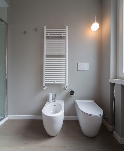 Nowoczesna łazienka z bidetem i toaletą w minimalistycznym stylu, grzejnik drabinkowy na ścianie, oświetlenie punktowe i kabina prysznicowa z boku.