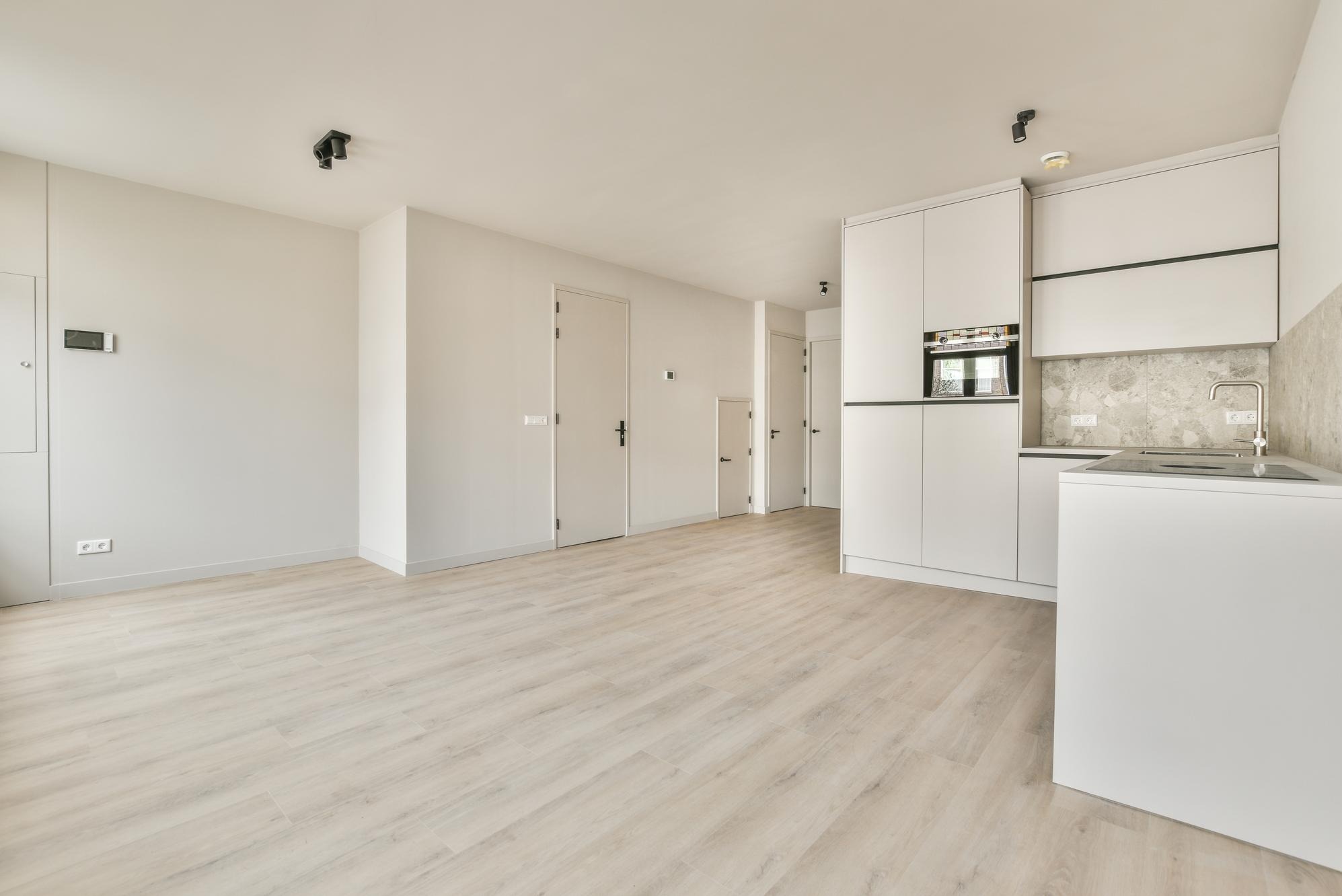 Wnętrze pustego, jasnego pomieszczenia z parkietem, białymi ścianami i zabudową kuchenną z szarym marmurkowym backsplashiem oraz czarnymi reflektorami sufitowymi.