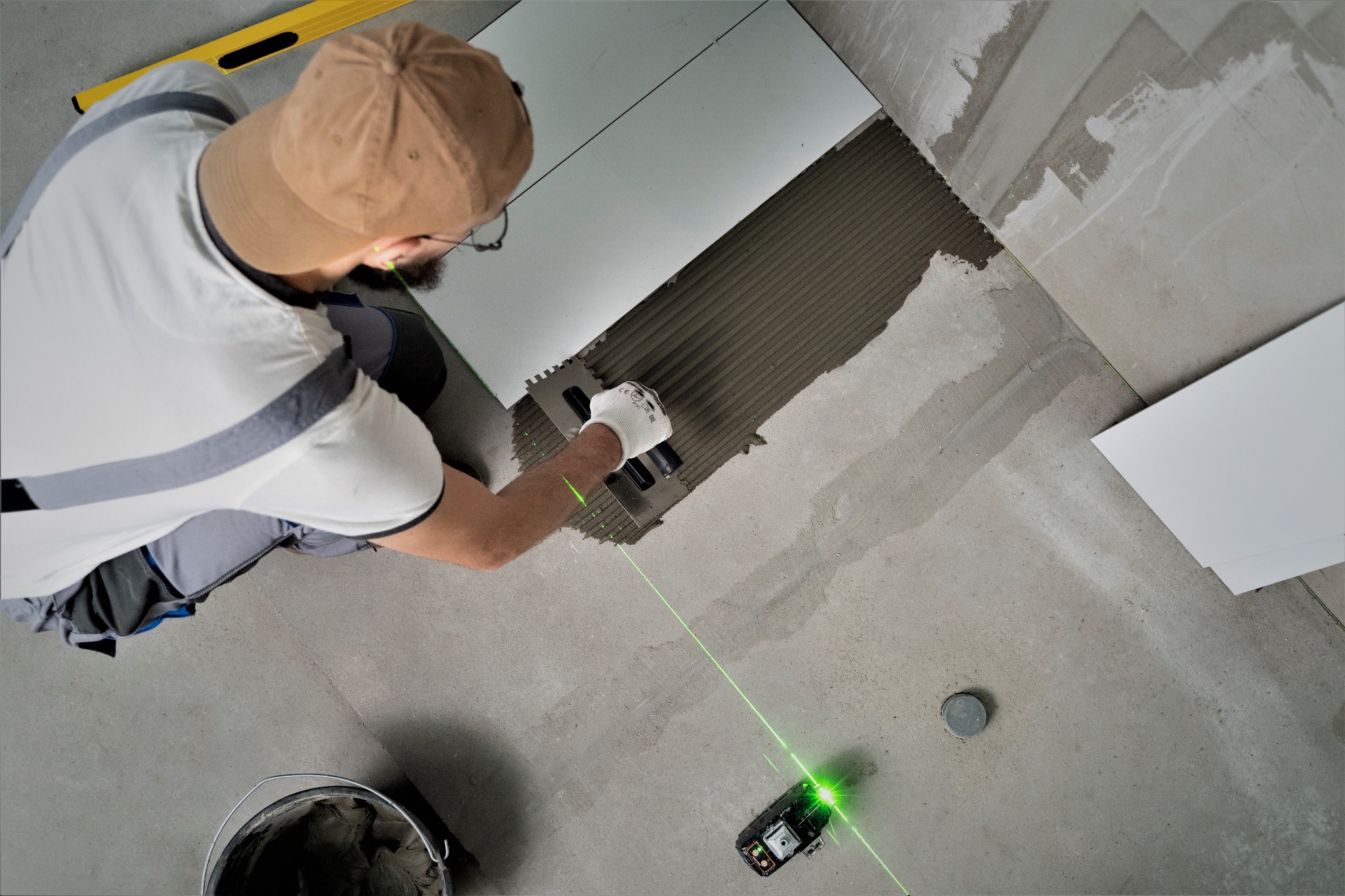 Układanie jasnych płytek podłogowych z użyciem poziomicy laserowej, widoczna paca z klejem oraz wiadro z zaprawą.