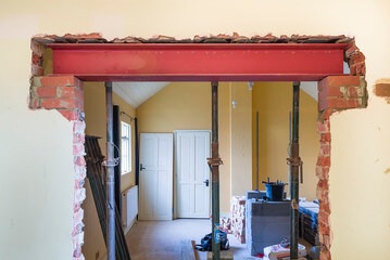 Montaż stalowej belki nośnej w otworze ściany z widocznymi fragmentami cegieł i wspornikami, wnętrze z białymi drzwiami i żółtą ścianą w tle.