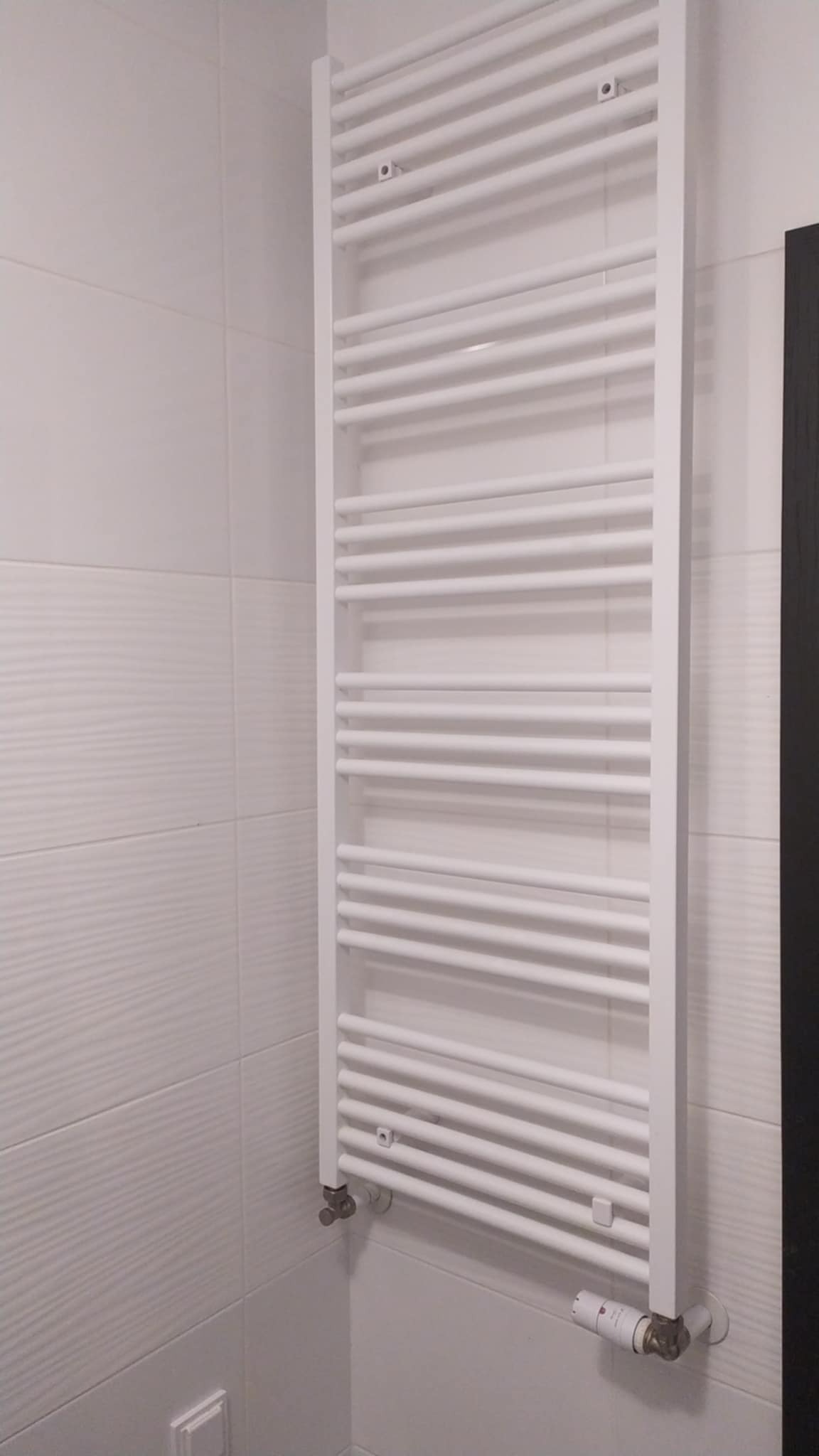 Biały, drabinkowy grzejnik łazienkowy na tle białych, strukturalnych płytek. Widoczny termostat i zawory. Fragment czarnej szafki po prawej stronie.