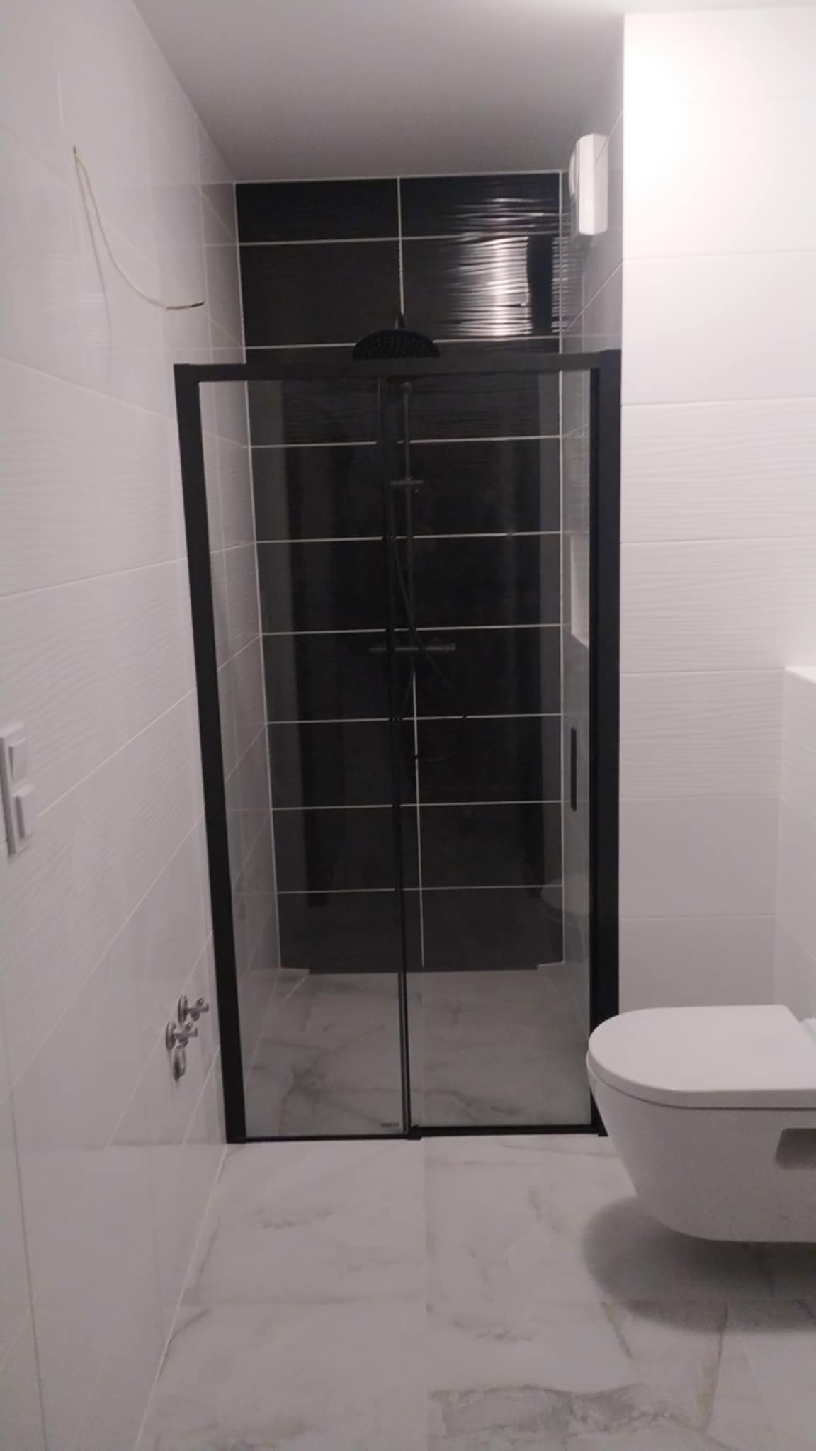 Nowoczesna łazienka z kabiną prysznicową z czarnymi ramami i deszczownicą, białe płytki imitujące drewno na ścianach, marmurowe płytki na podłodze, wisząca toaleta.