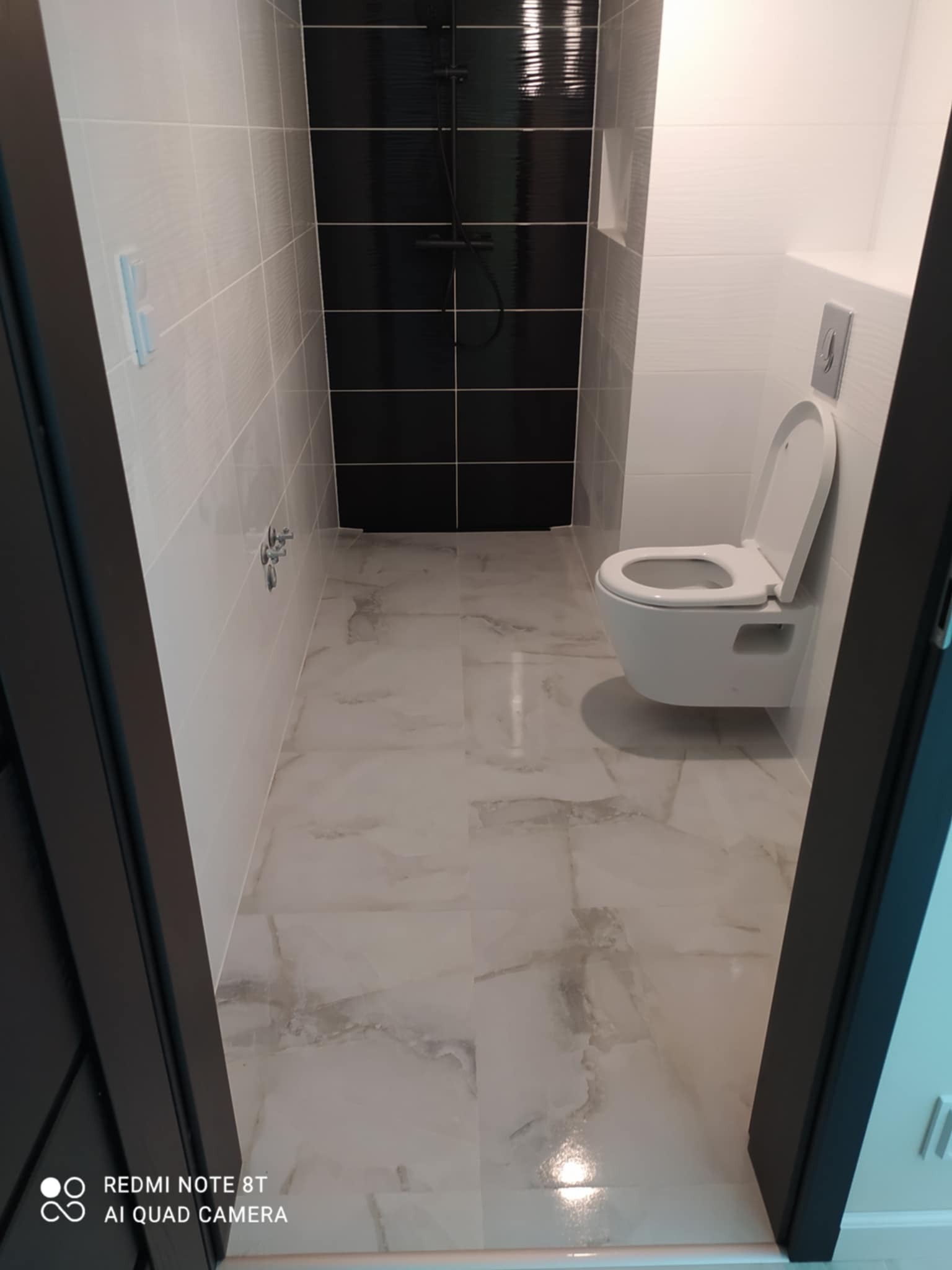 Nowoczesna łazienka z podwieszaną toaletą, białymi płytkami ściennymi z subtelną fakturą i czarnymi płytkami w strefie prysznica, widok przez czarną framugę drzwi.