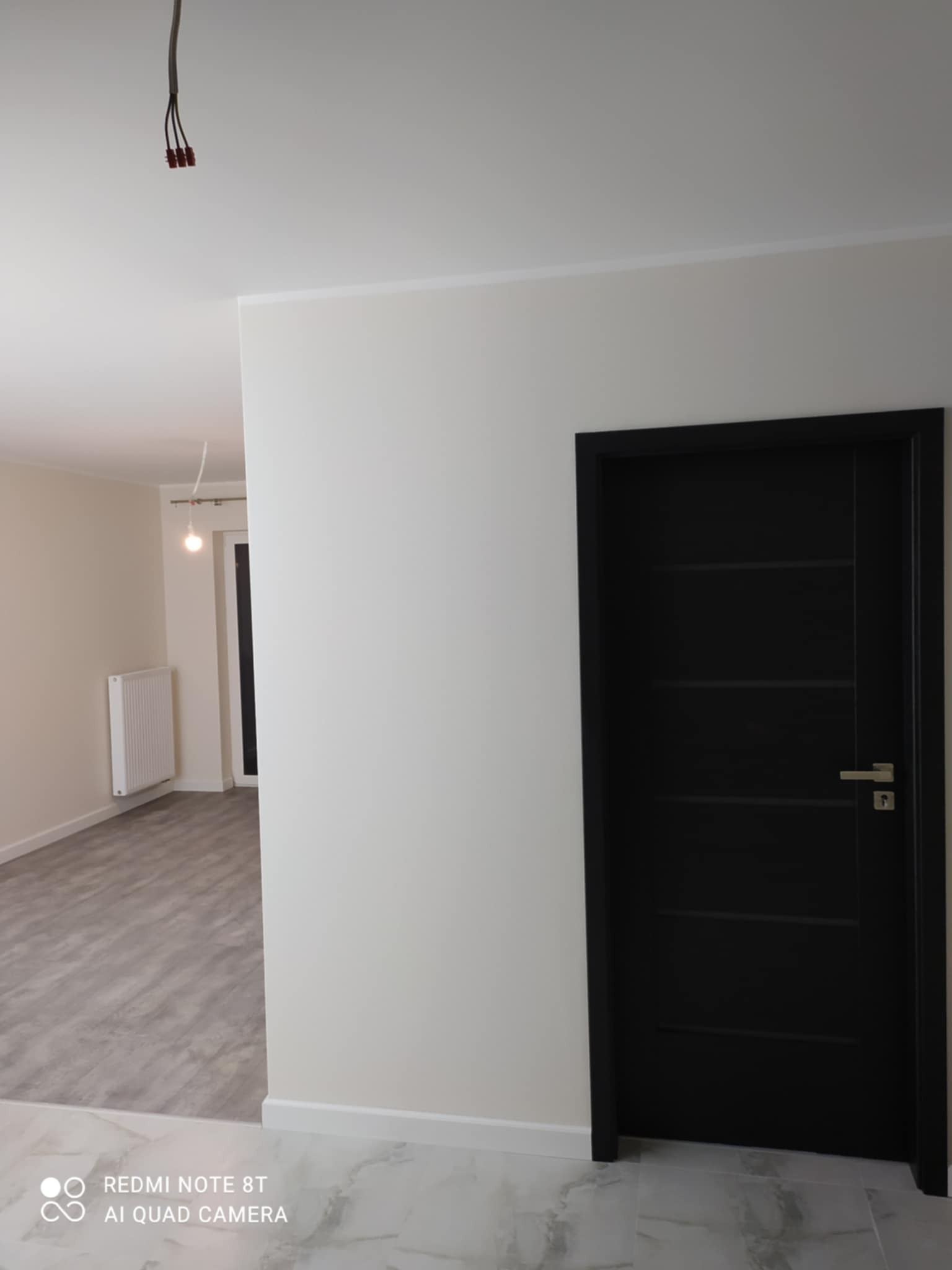 Wnętrze po remoncie: białe ściany, szara podłoga imitująca drewno, czarne drzwi wewnętrzne z poziomymi frezami, widoczne kable wystające z sufitu.