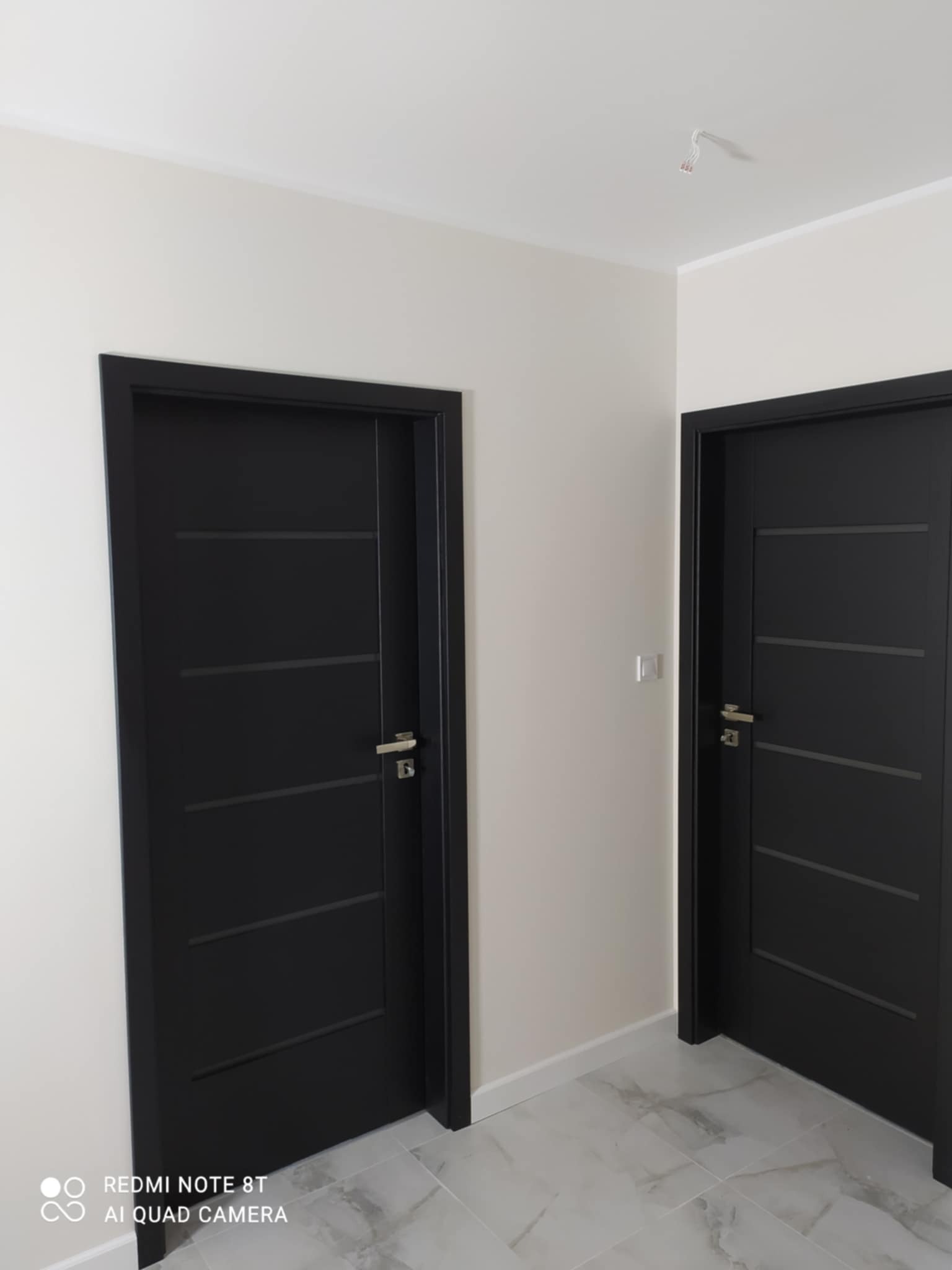 Dwa czarne, nowoczesne drzwi wewnętrzne z poziomymi frezami, zamontowane w jasnym pomieszczeniu z marmurową podłogą i białymi ścianami. Widoczne wystające kable ze ściany w miejscu lampy.