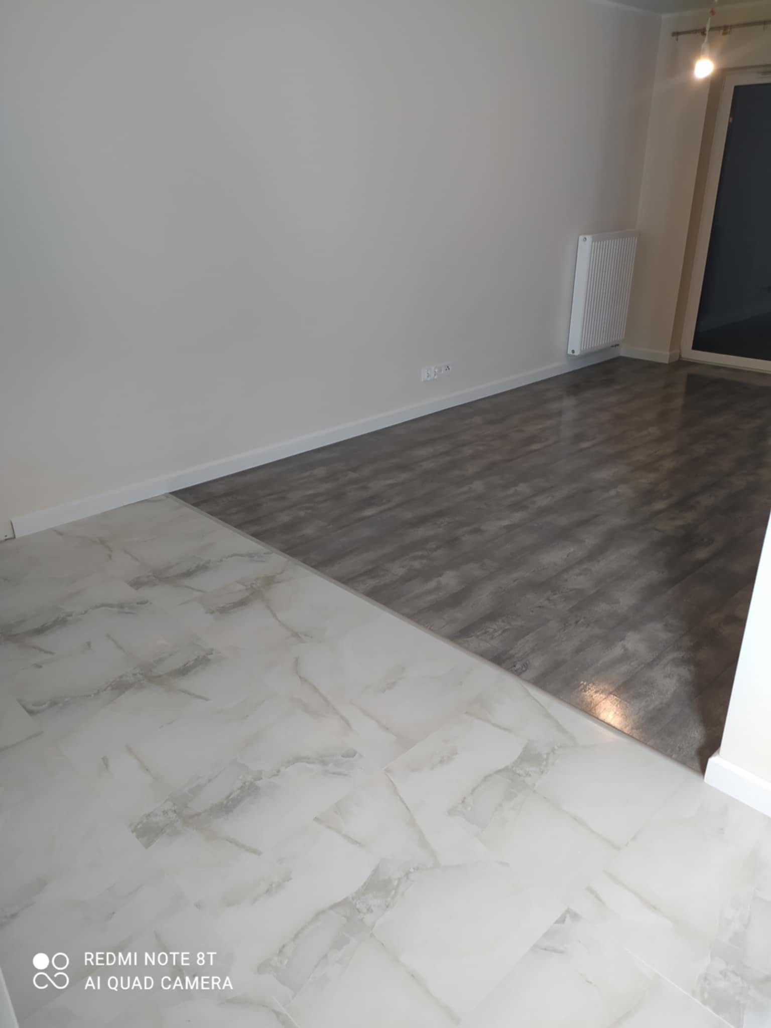 Połączenie dwóch rodzajów podłóg: białe płytki imitujące marmur i szara, drewnopodobna podłoga w pustym pokoju z białymi ścianami i grzejnikiem.
