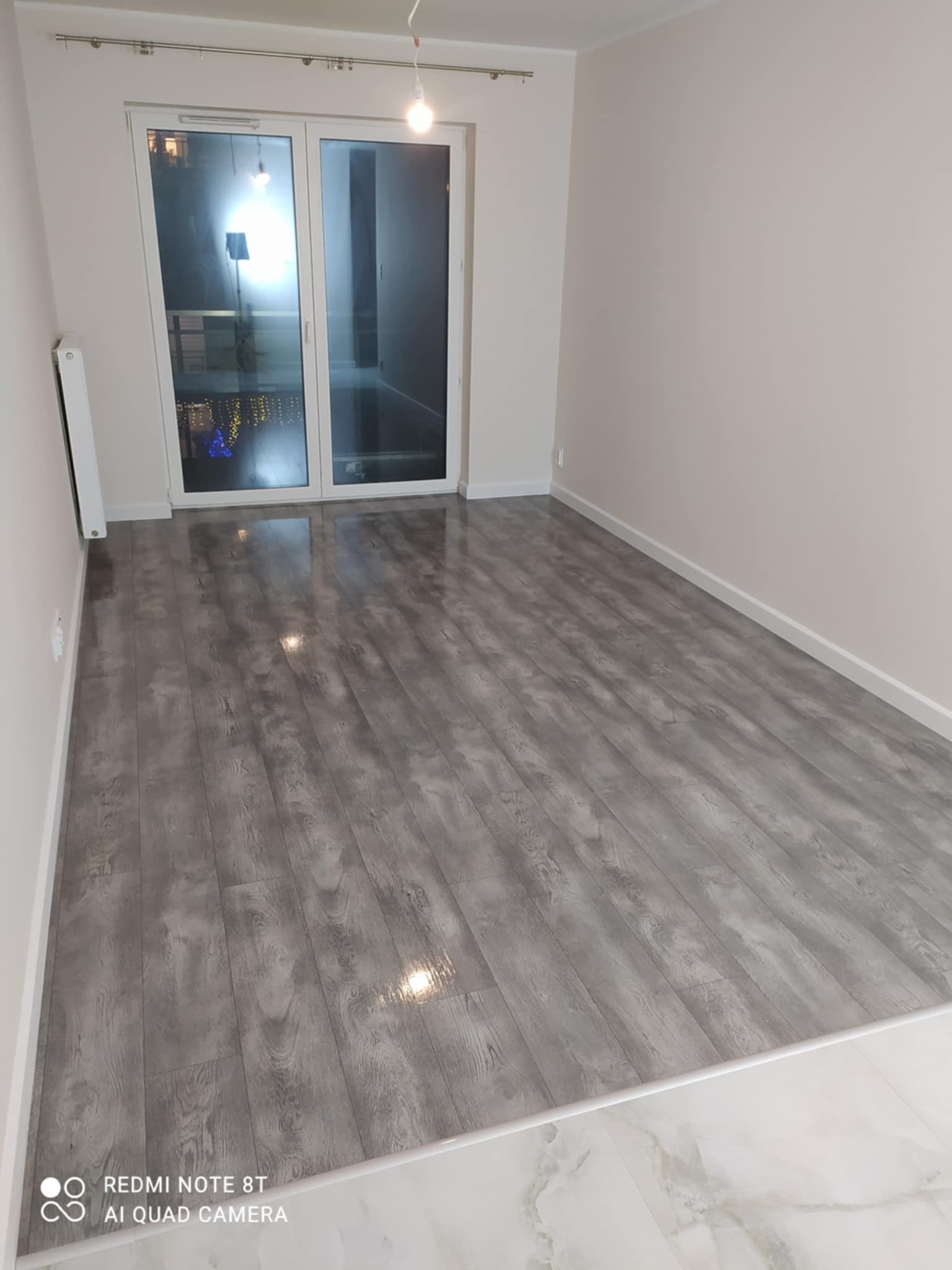 Pusty pokój po remoncie z szarą podłogą imitującą drewno i białymi ścianami, widoczne okno balkonowe z widokiem na oświetlone miasto nocą.