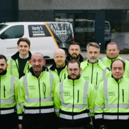 Grupa pracowników Hardy's Industries w odblaskowych kurtkach przed budynkiem firmy i firmowymi samochodami dostawczymi z logo.