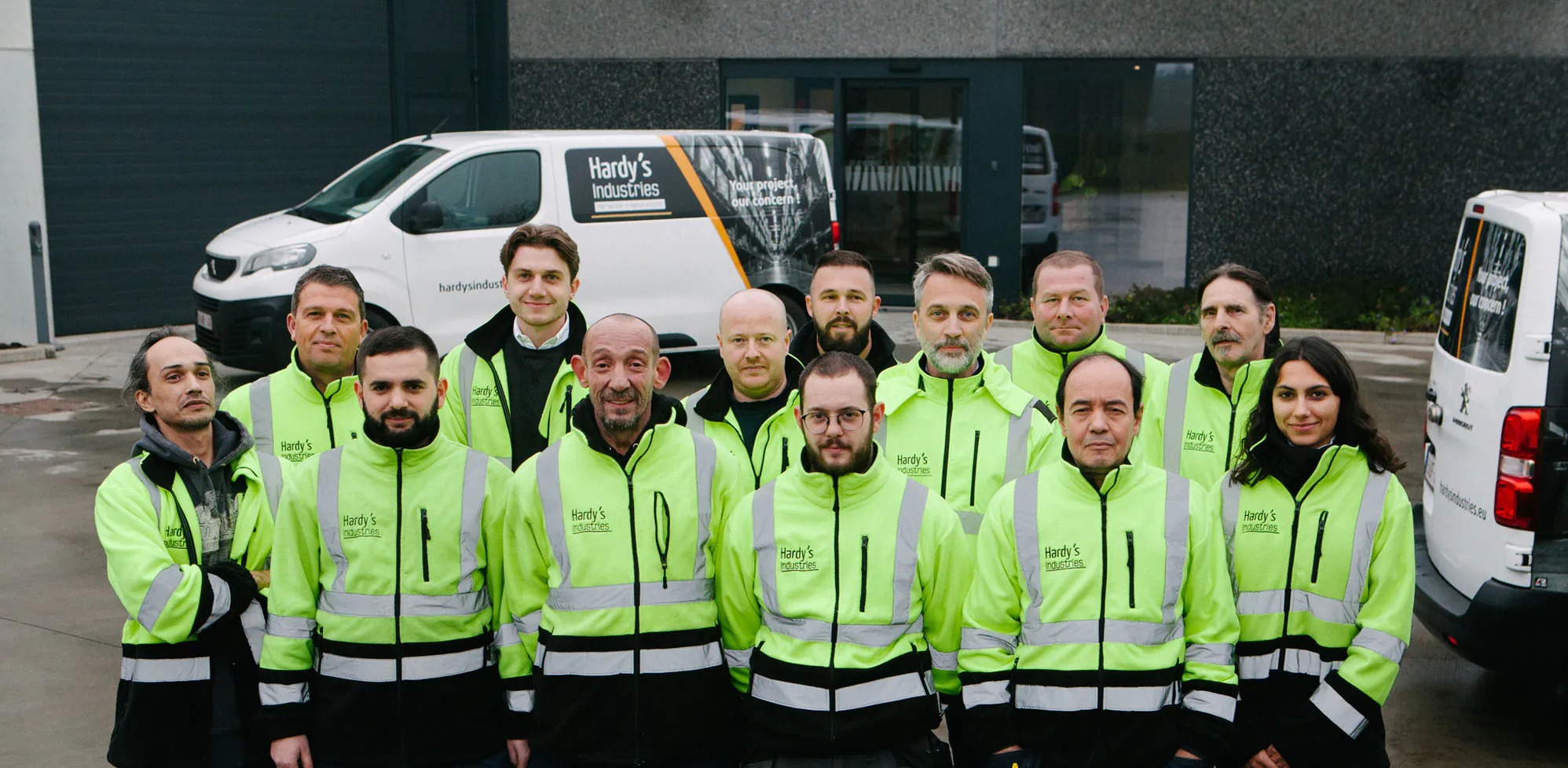 Grupa pracowników Hardy's Industries w odblaskowych kurtkach przed budynkiem firmy i firmowymi samochodami dostawczymi z logo.
